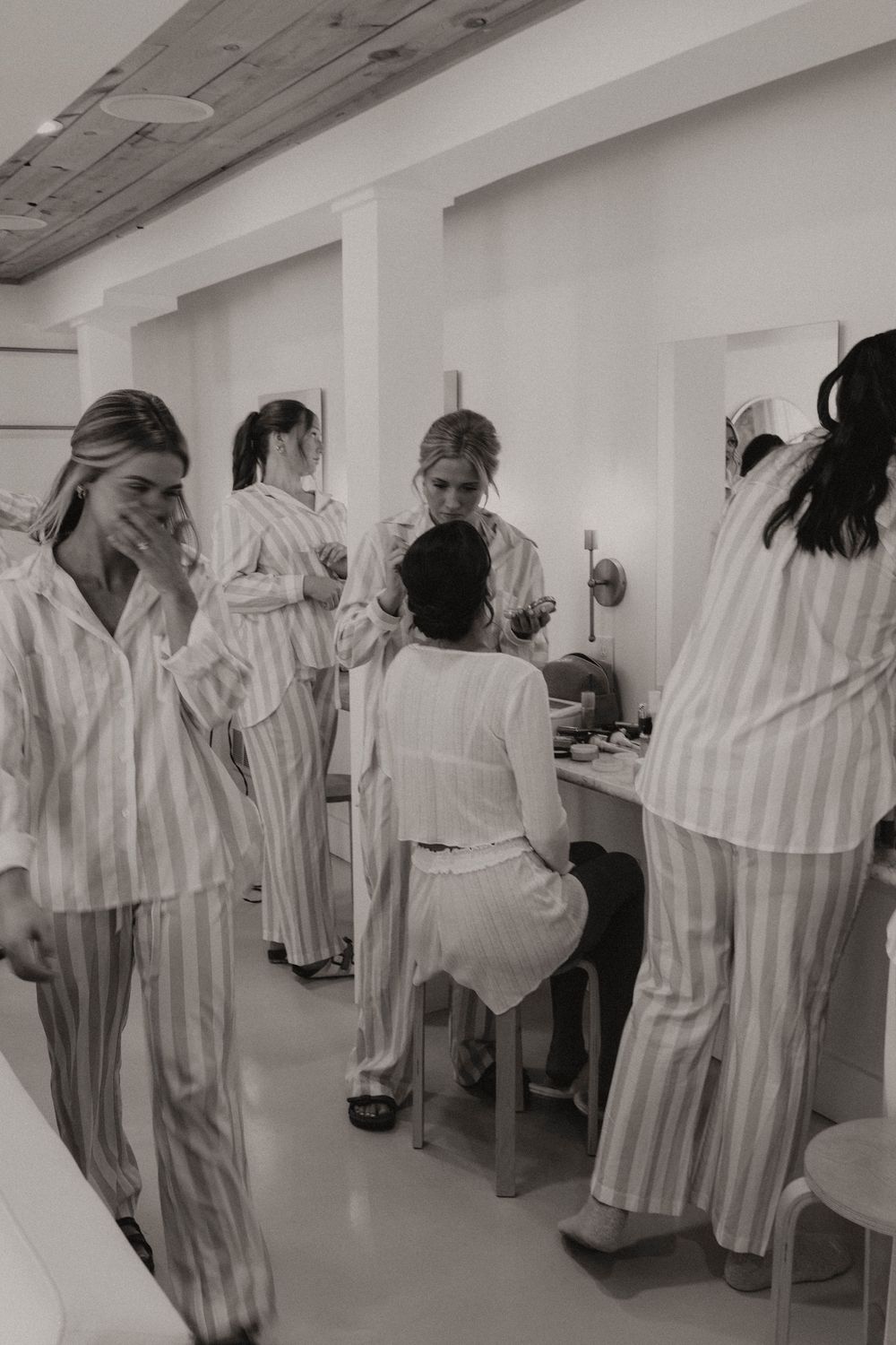 A group in matching striped pajamas gather around a makeup vanity table during wedding preparations.