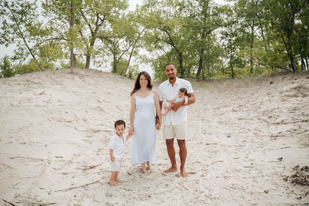 A family wearing white outfits stands together on sandy dunes surrounded by trees on a sunny day.