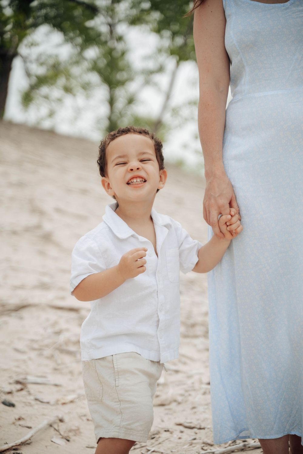 A child in white shirt and khaki shorts holds hands with someone in a light blue dress while walking on sand.