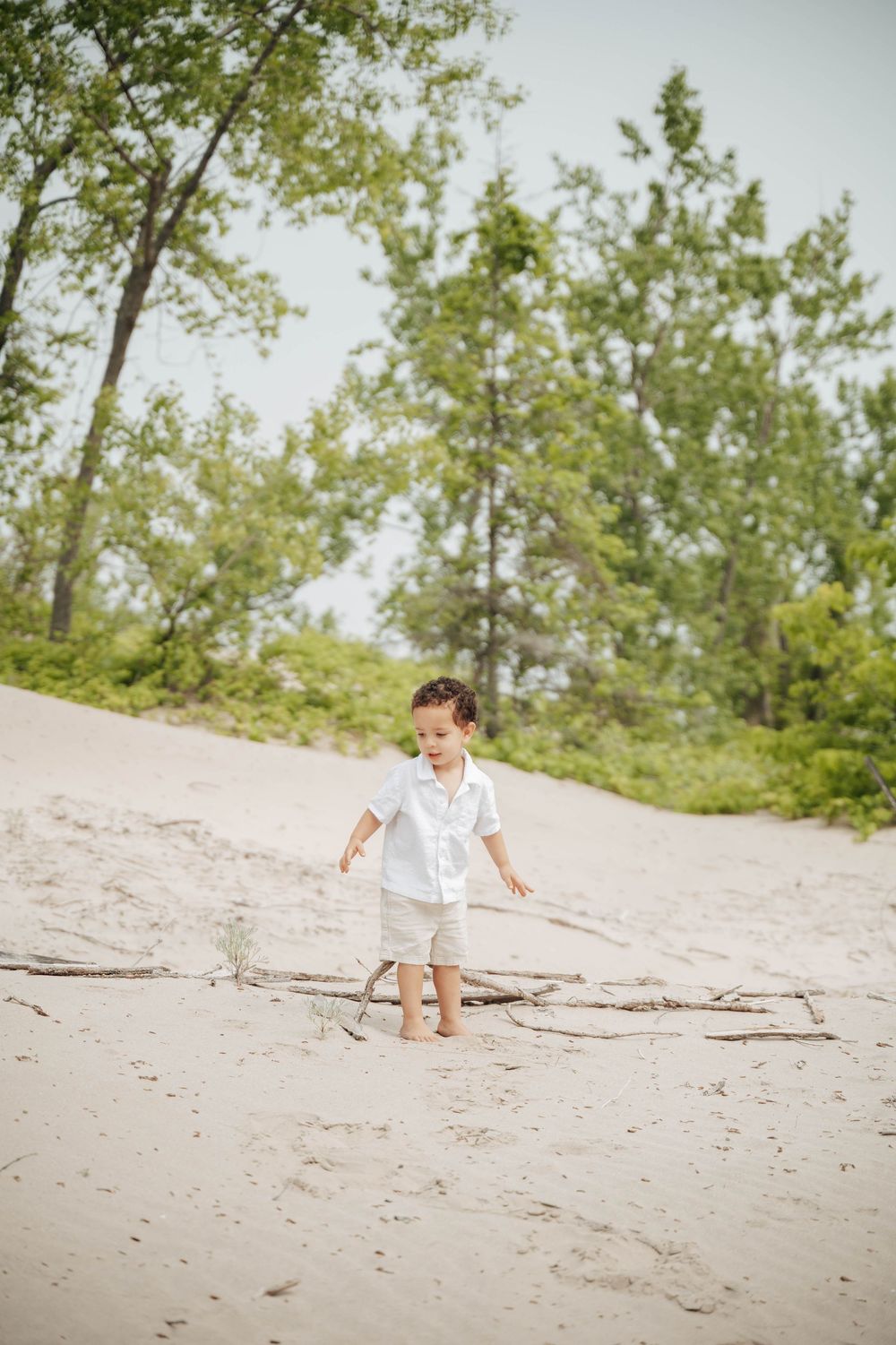 A child in white clothing plays on a sandy beach surrounded by tall green trees on a sunny day.