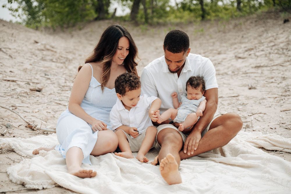 A family sits together on a blanket at the sandy beach wearing coordinated white outfits on a sunny day.