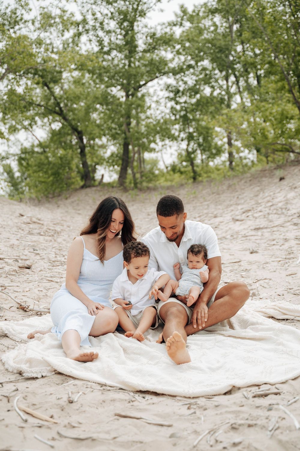 A family wearing white outfits sits together on a blanket at a sandy beach near trees.