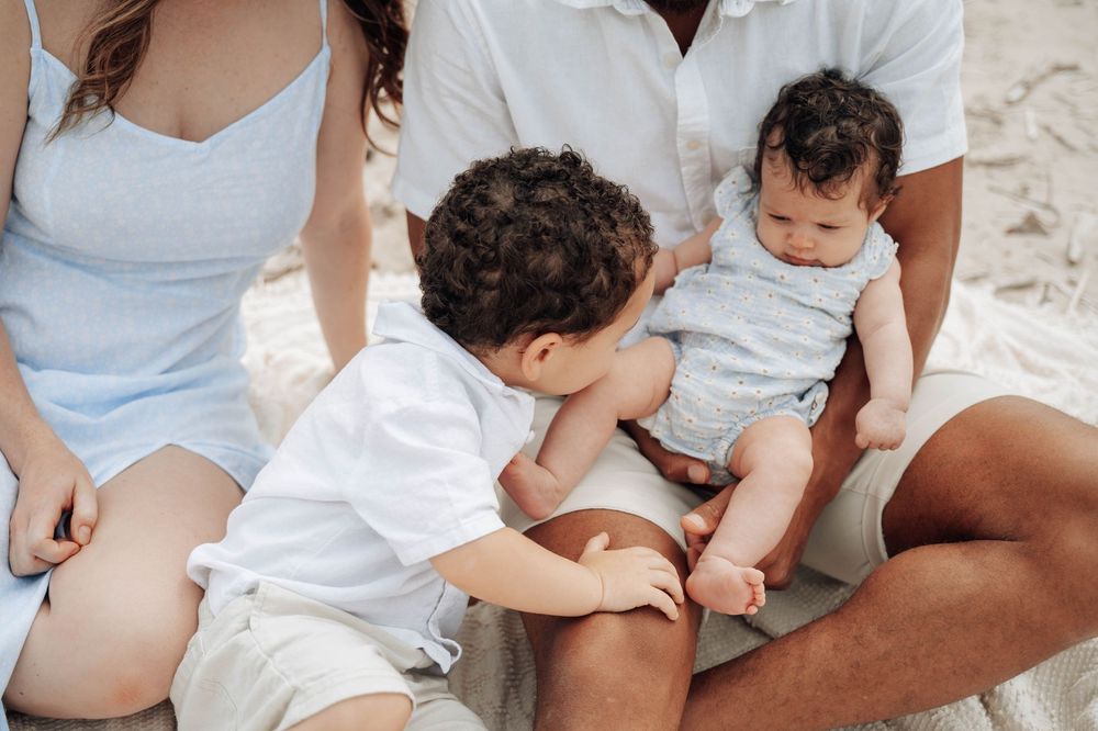 Two siblings interact sweetly while sitting with their family, wearing coordinated white and light blue outfits.