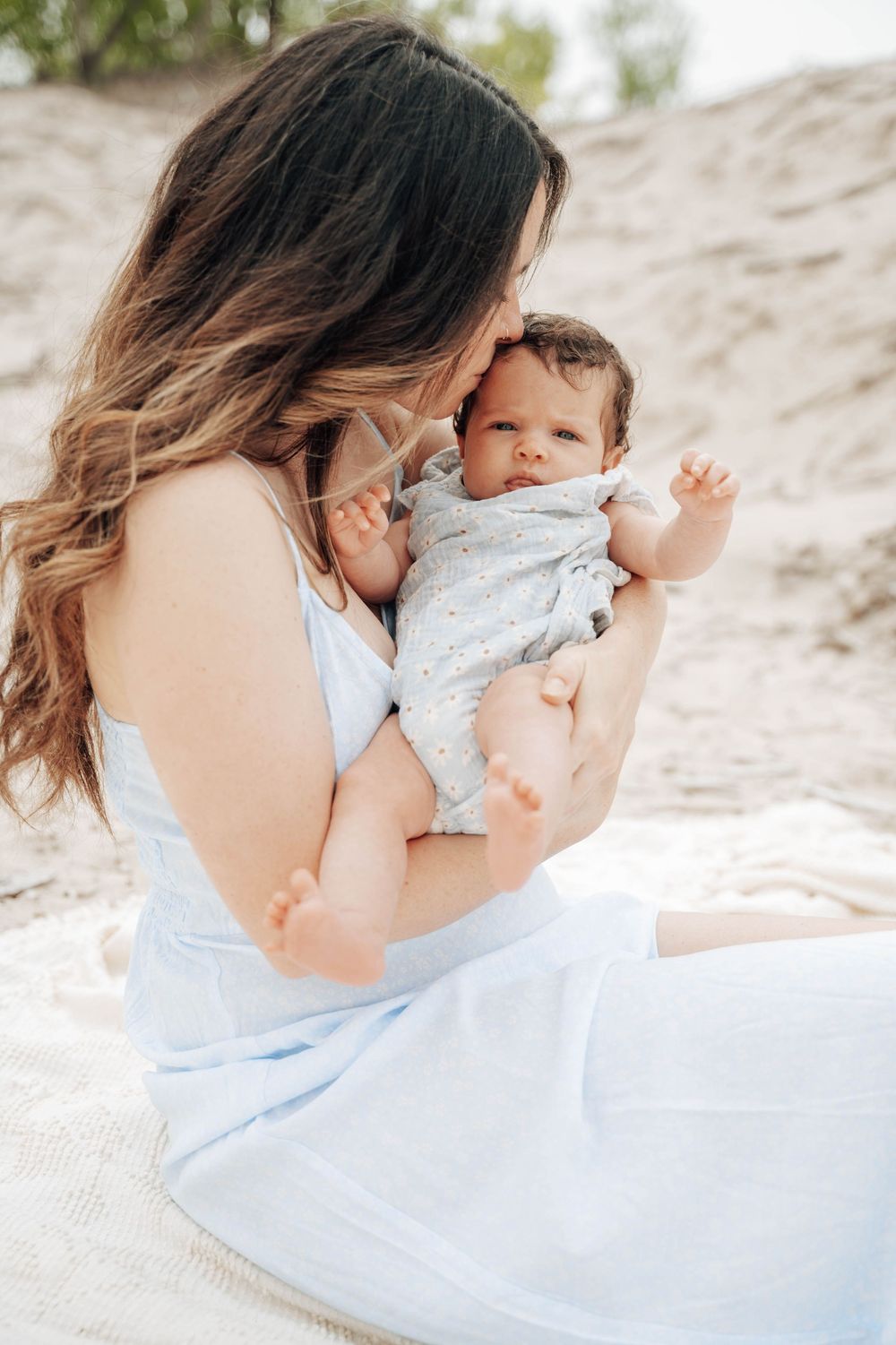 A tender moment between mother and infant baby on a sandy beach, wearing light blue and white clothing.