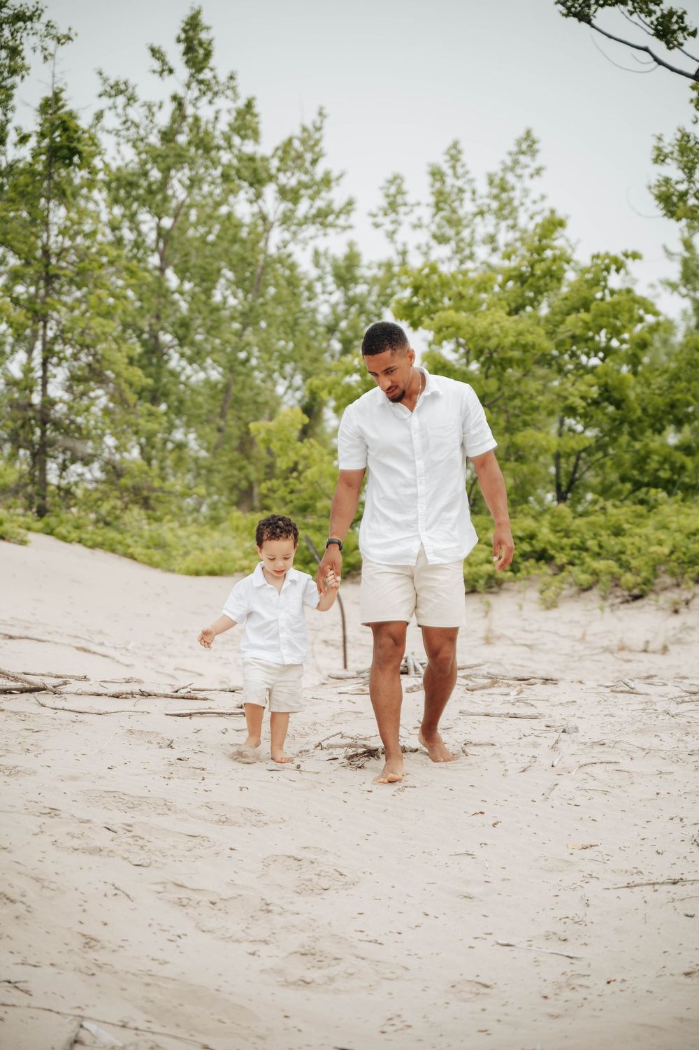 Two people in matching white outfits walk barefoot together on a sandy beach path bordered by green trees.