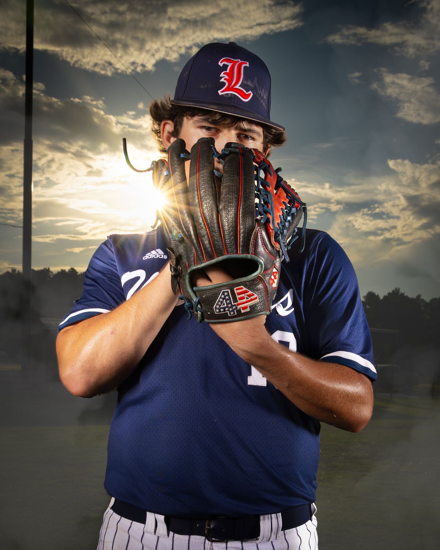 Baseball player in navy blue uniform holds glove over face as dramatic sunset sky glows behind them.