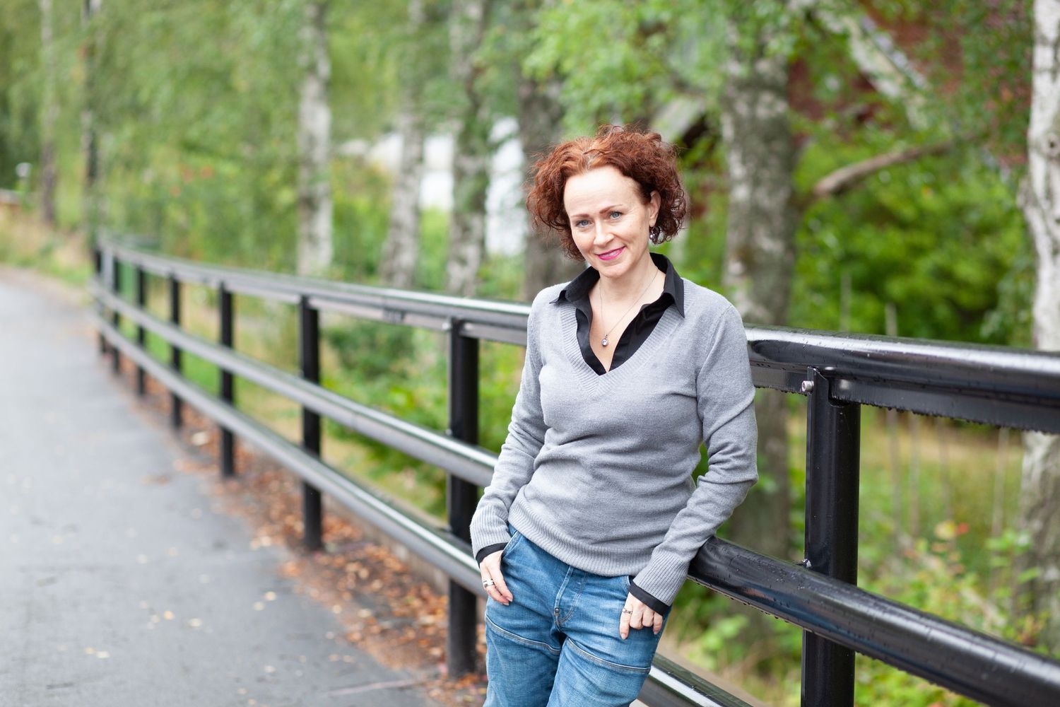 A person in a grey sweater and jeans stands smiling along a wooden railing on a path surrounded by green trees.