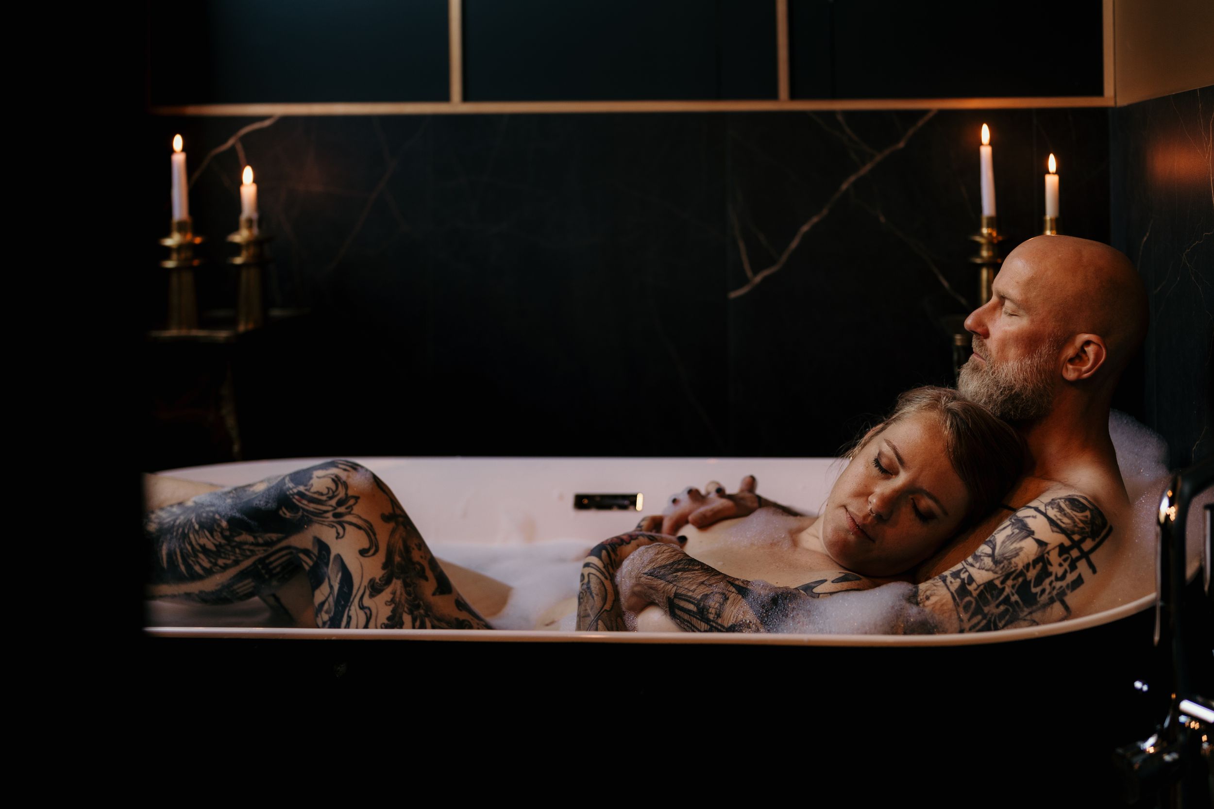 A candlelit bathtub scene with two tattooed people relaxing in intimate atmospheric lighting.