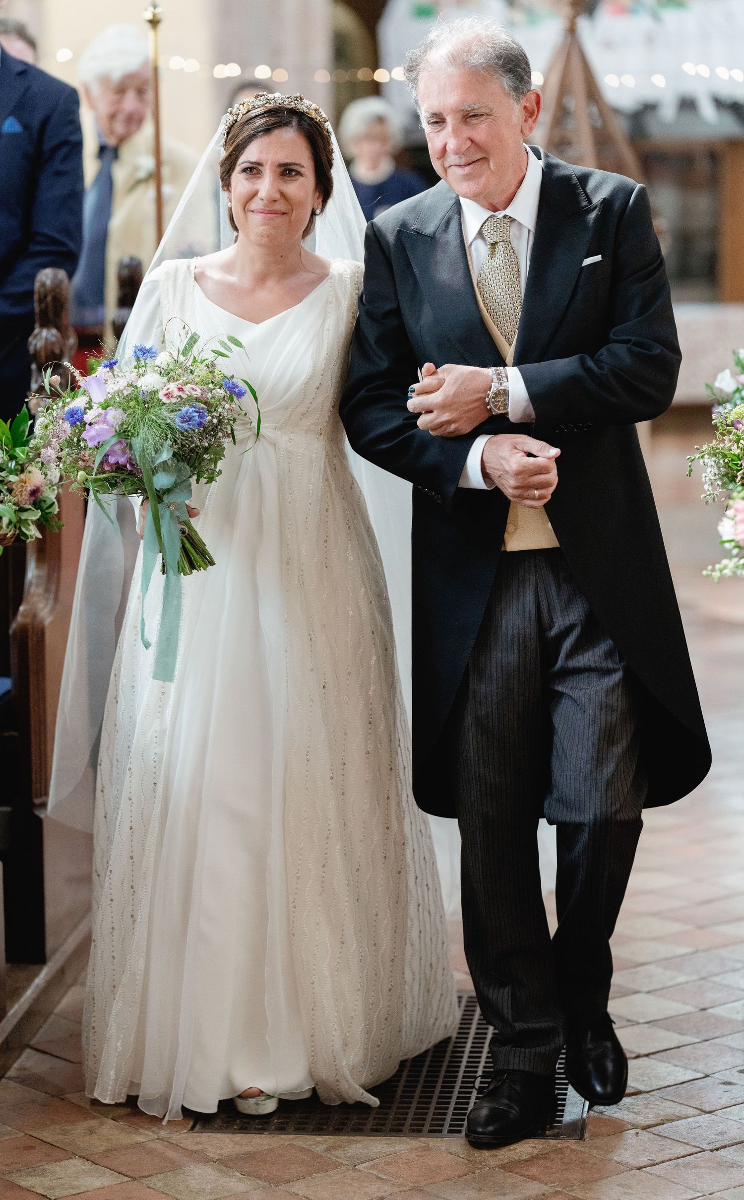 A bride in white wedding gown holds a purple and blue wildflower bouquet while walking arm in arm with someone in formal attire.