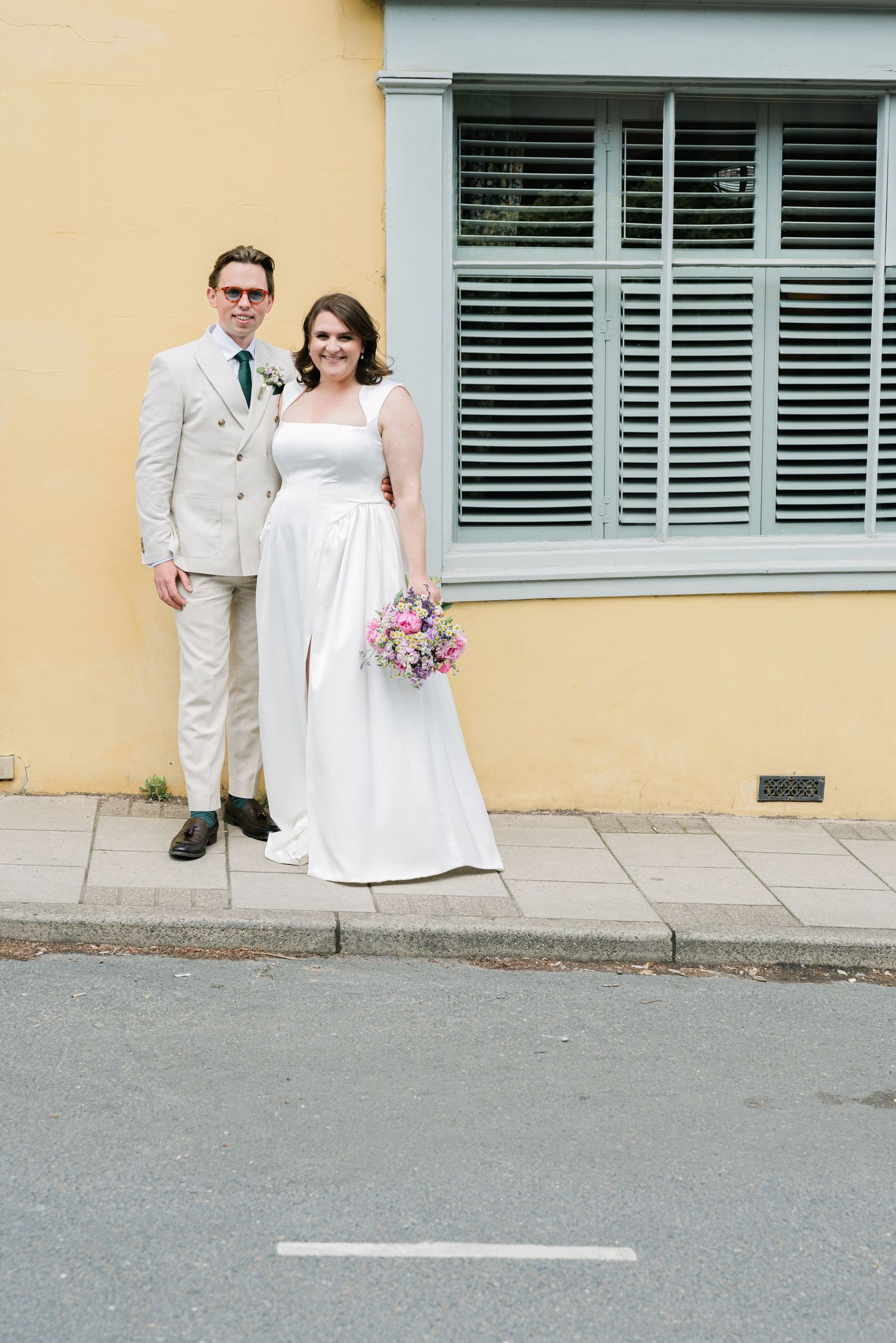 A couple in wedding attire poses together on a sidewalk against a yellow wall with shuttered window.