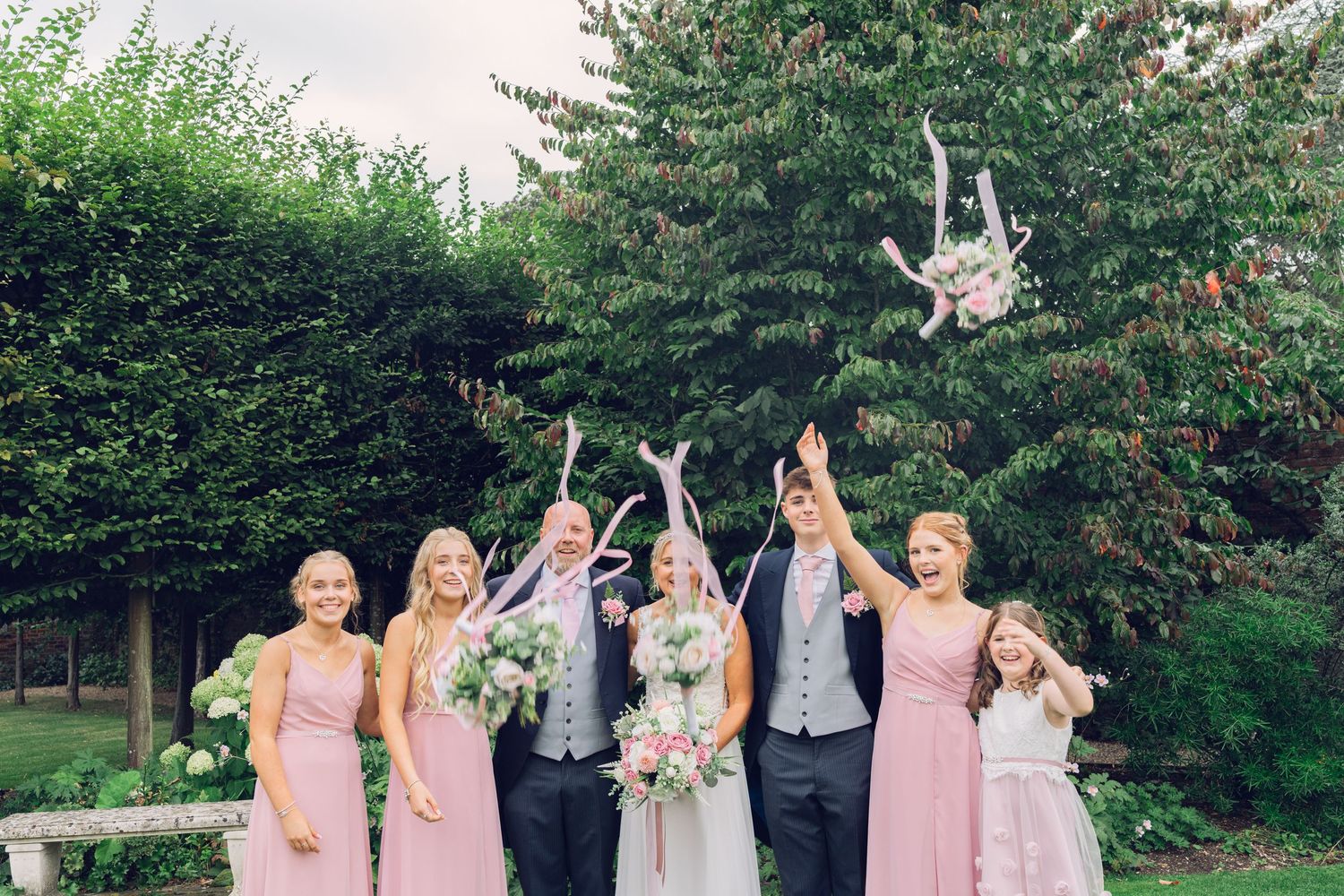 Wedding party celebrating by tossing a bouquet in the air in front of green hedges and trees.