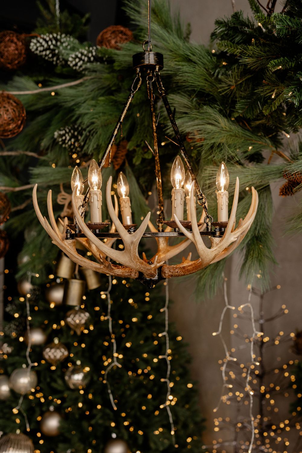 Rustic antler chandelier illuminates Christmas tree with warm candle-style lights and festive pinecone decorations.