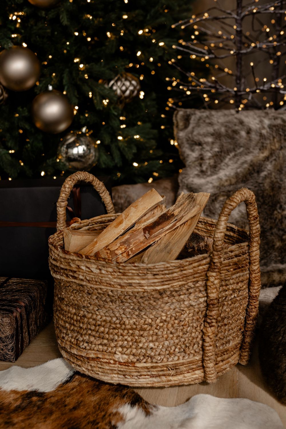 A woven basket filled with firewood sits beside a Christmas tree decorated with silver ornaments and twinkling lights.