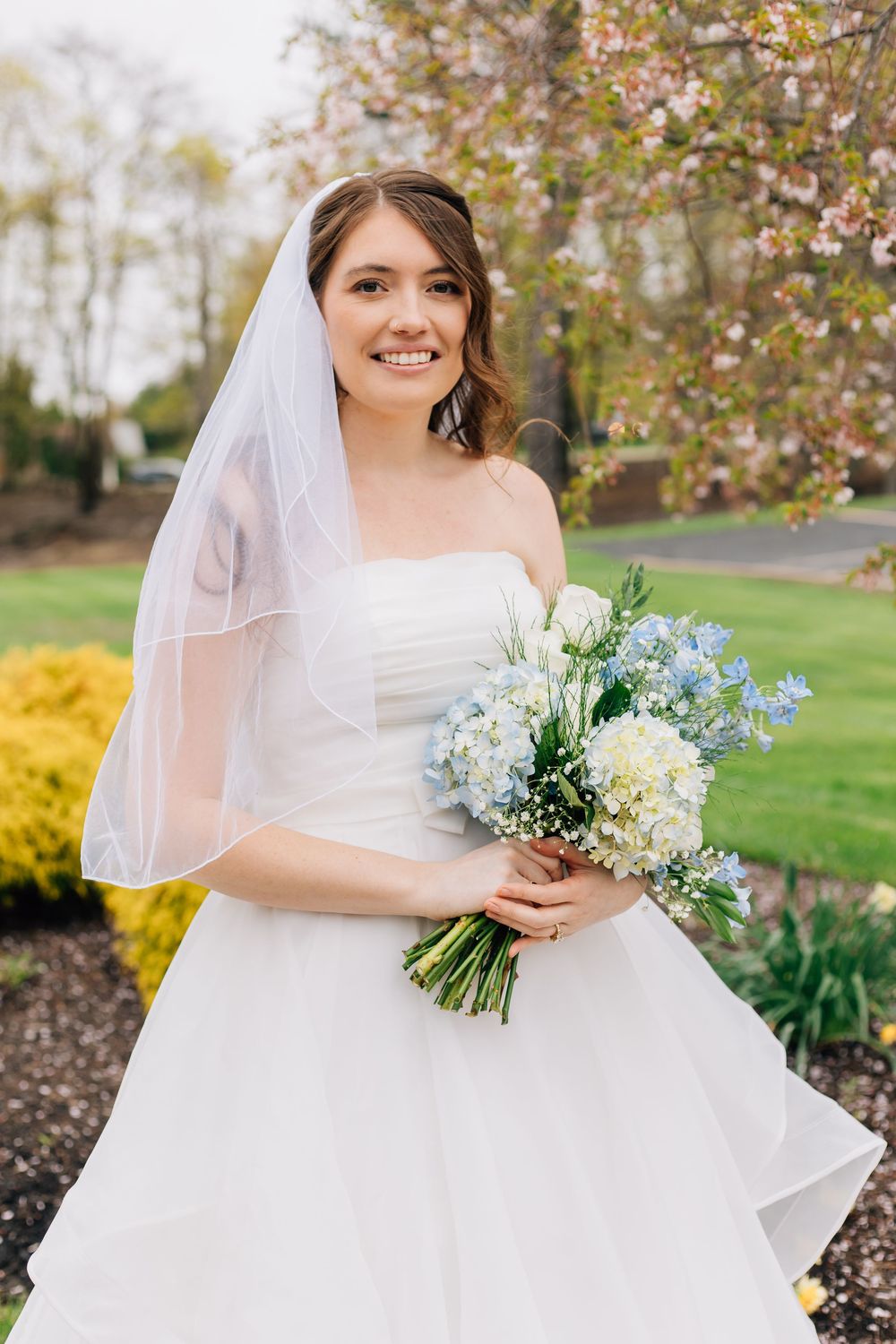 Bride in traditional white gown holds spring bouquet outdoors with cherry blossoms.