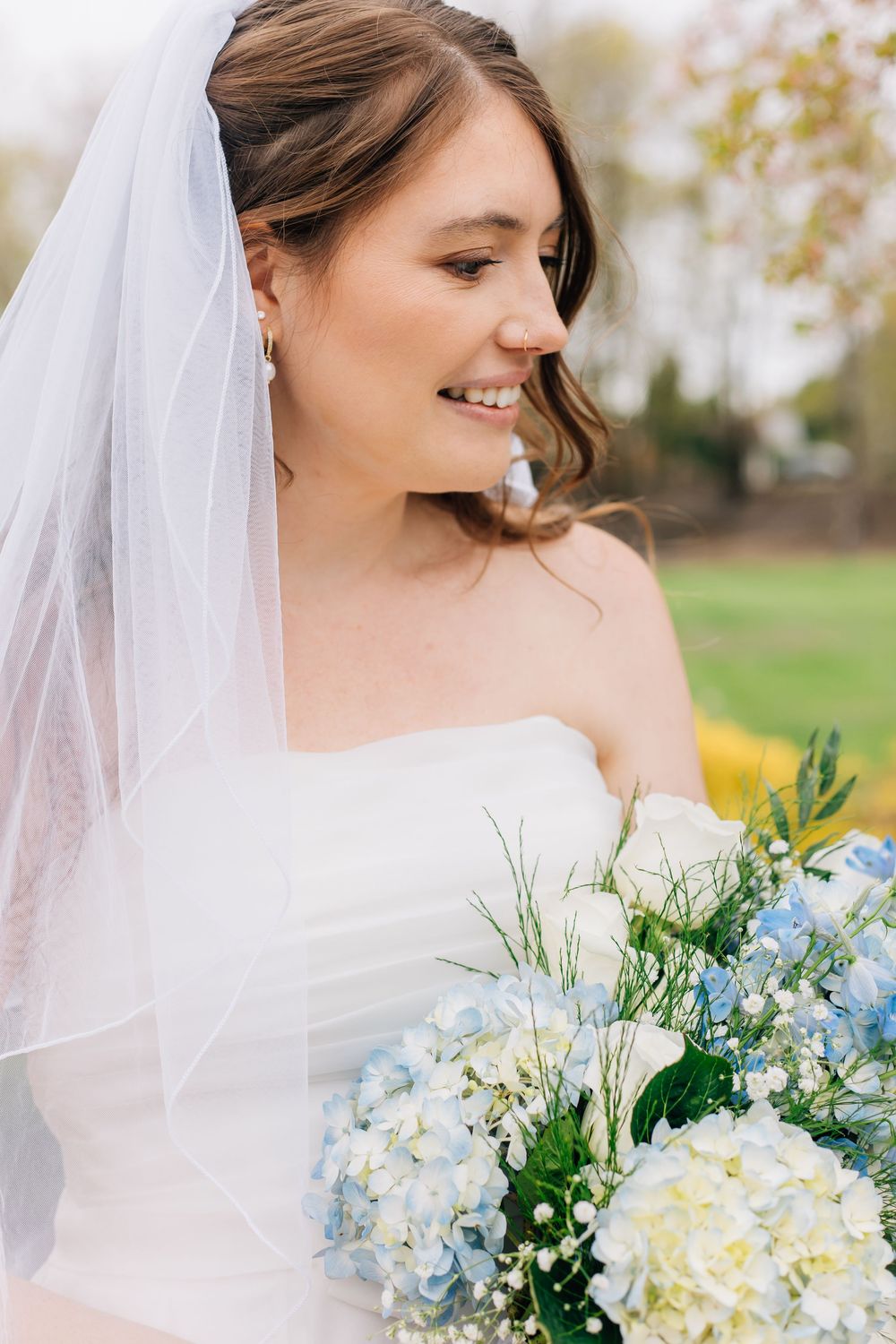 Bride wearing white dress and veil holds blue and white bouquet outdoors.