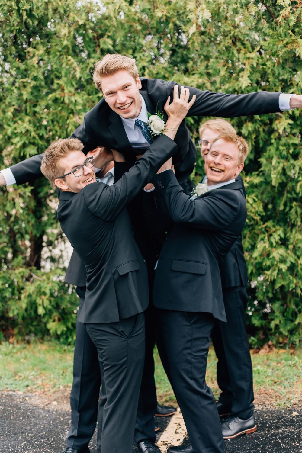 Groomsmen in dark suits playfully lifting someone during wedding photos.