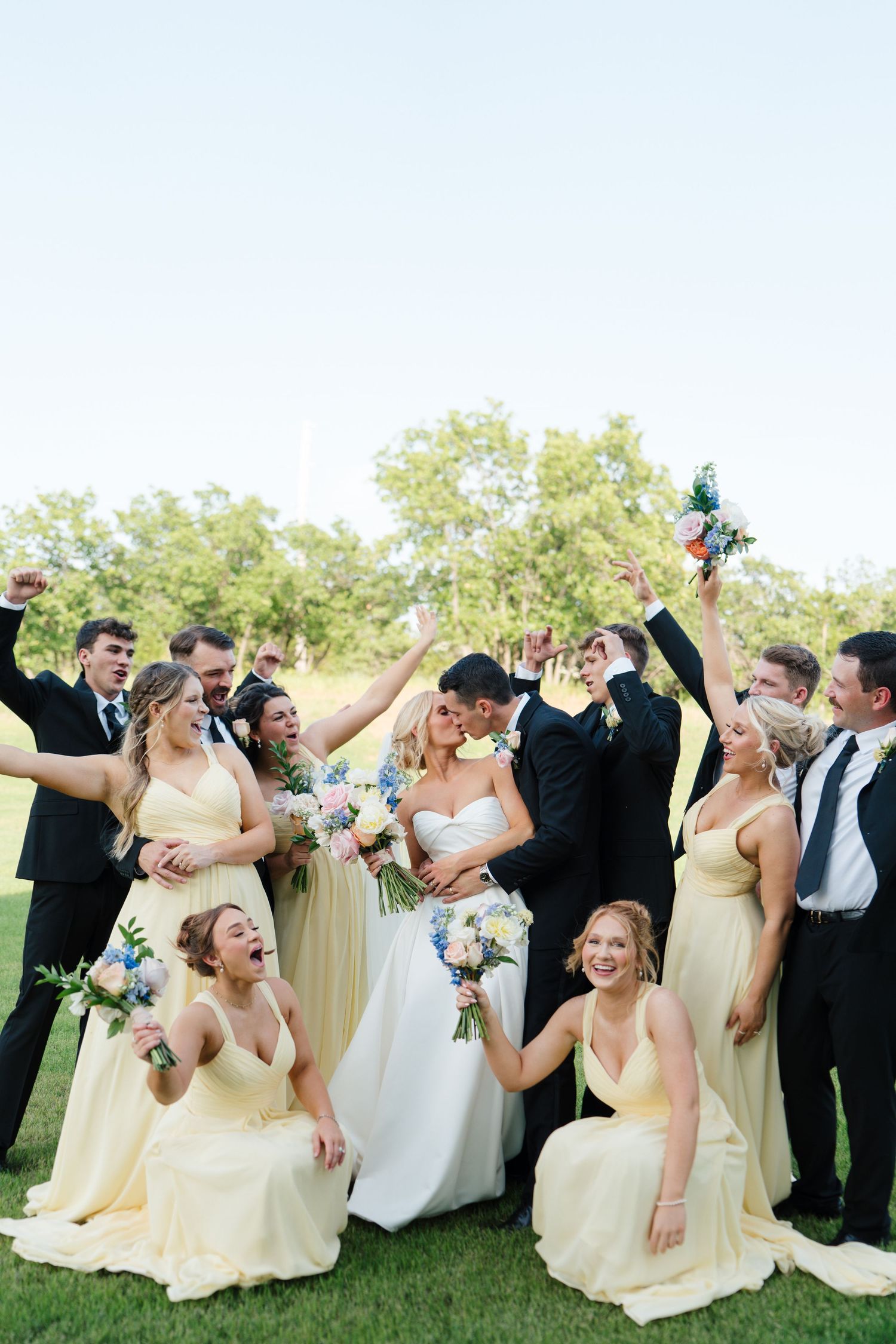 Wedding party in pale yellow bridesmaid dresses and black suits celebrates joyfully on a sunny lawn with bouquets.