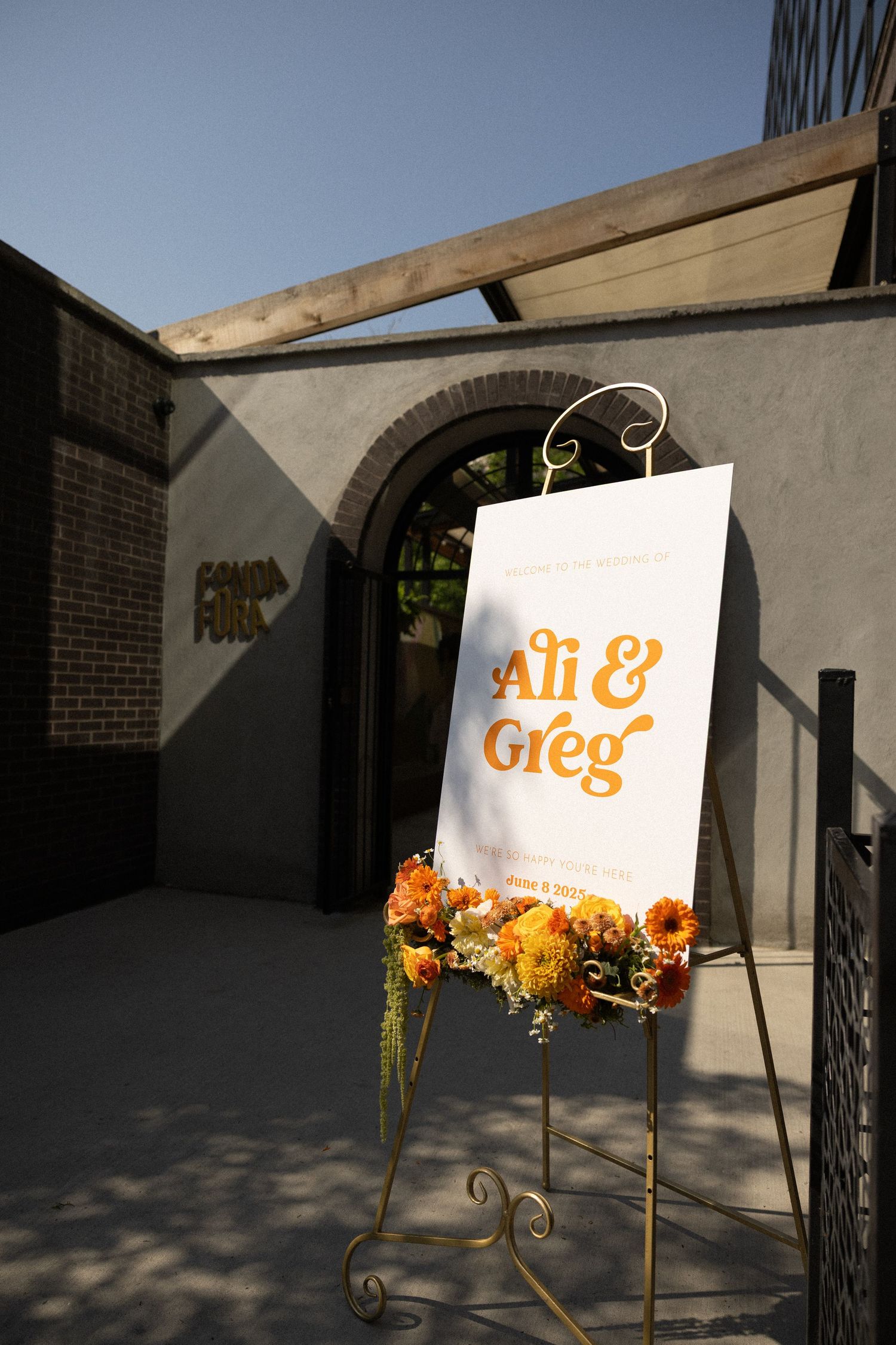 White paper gift bag with orange "Ali & Greg" text sits on ornate stand decorated with orange and yellow flowers.