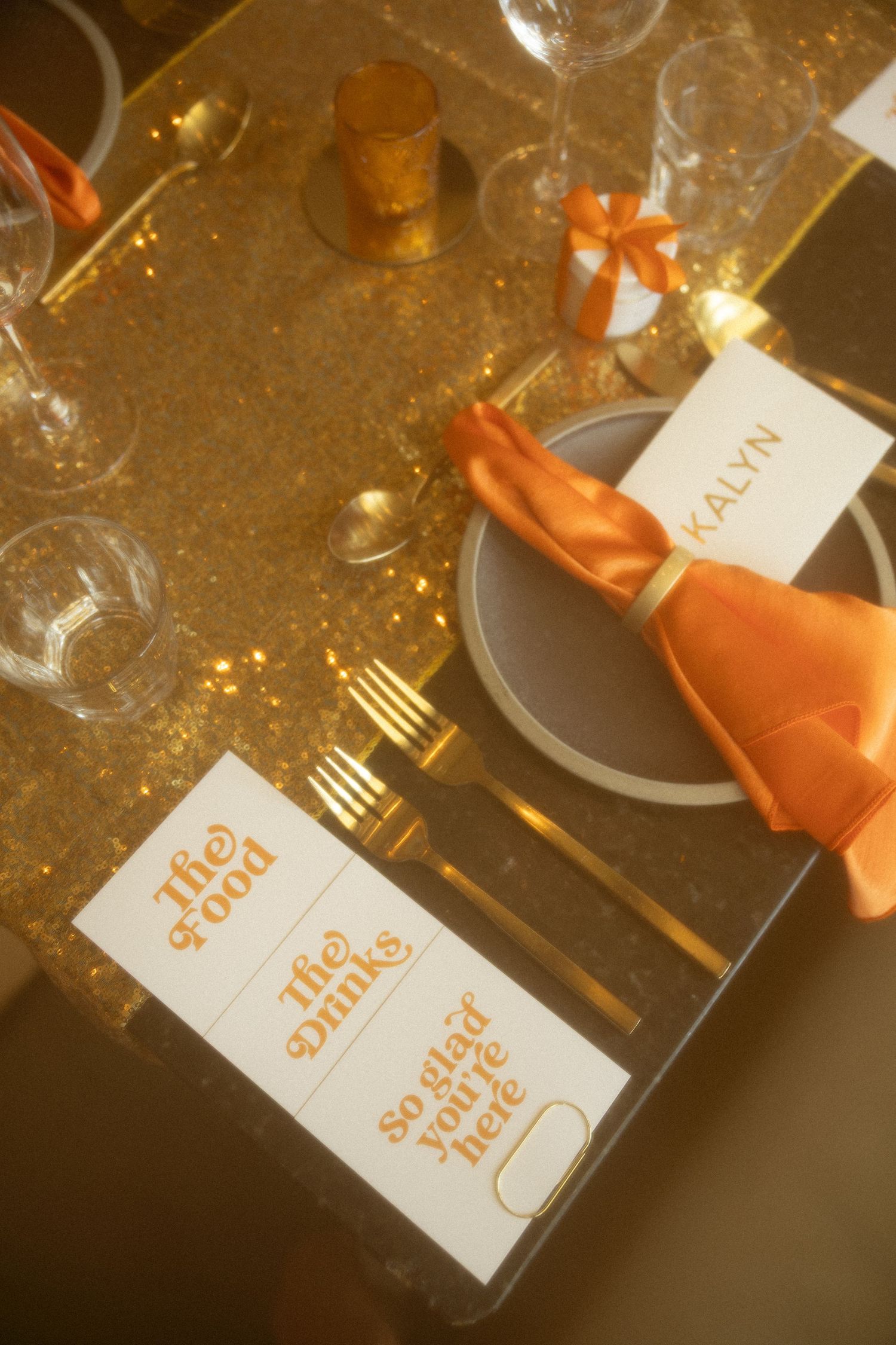 Wedding reception place settings with orange napkins and name cards on a golden table.
