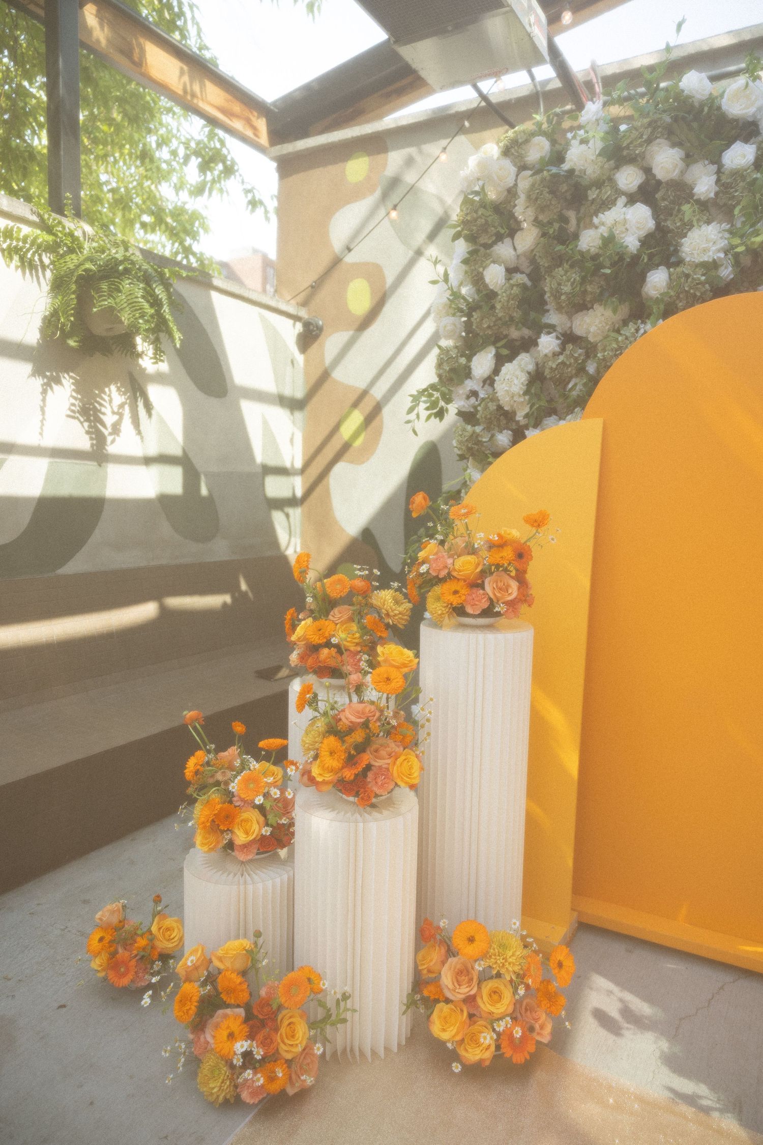 Minimalist wedding decor with white columns and orange floral arrangements against yellow curved wall.