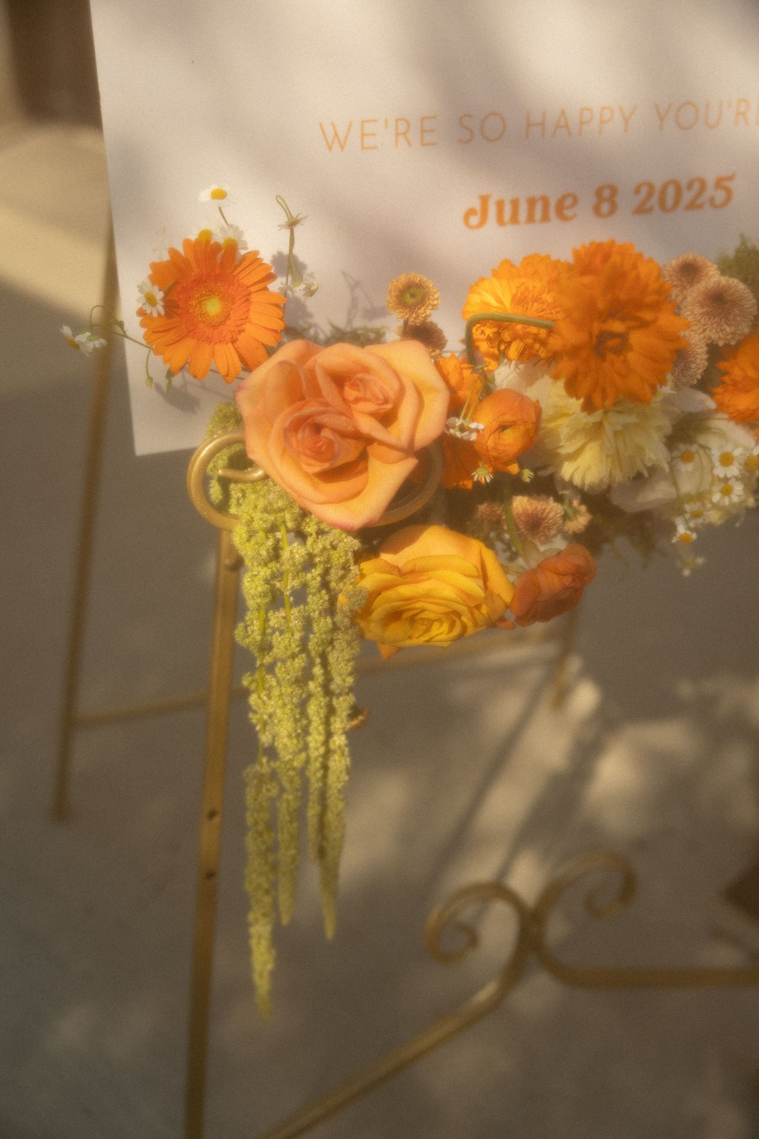A collection of orange roses and marigolds with trailing greenery arranged against a sunlit wall.