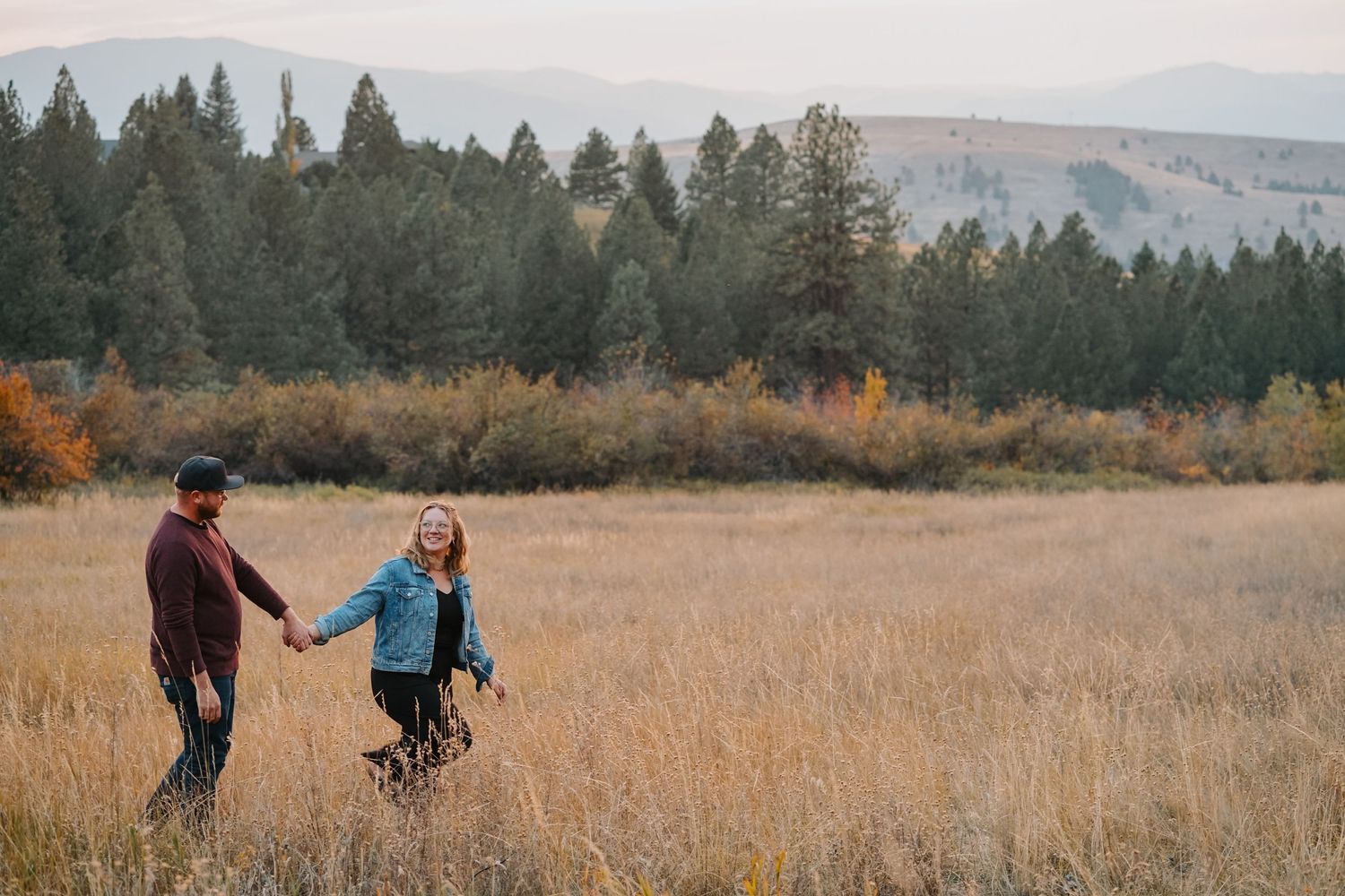 Couple joyfully dancing together in a golden field at sunset with evergreen trees in the background.