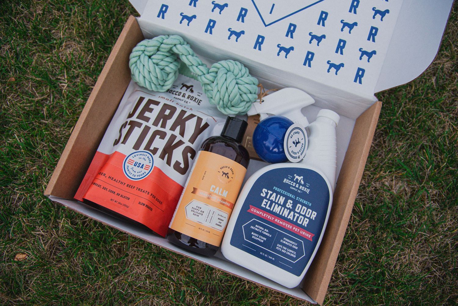 Rocco & Roxy pet care and treat box with jerky sticks, stain remover, and a rope toy on grass. Product photography