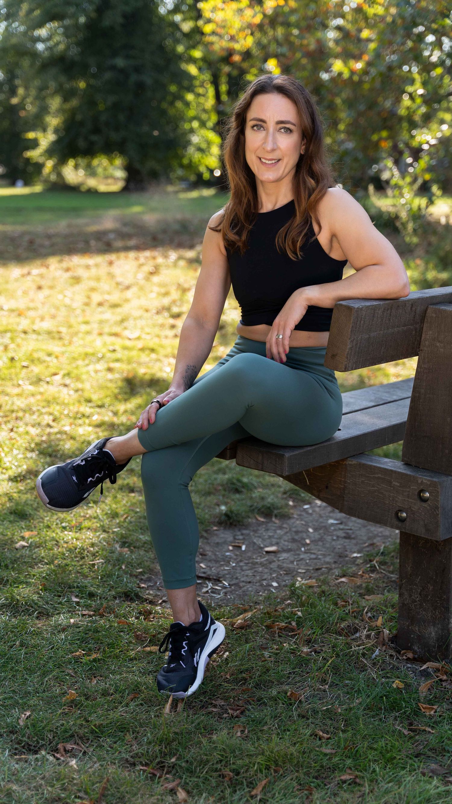 Female entrepreneur sits on a park bench on a personal branding photoshoot in West London. She's a PT and is wearing workout clothes.