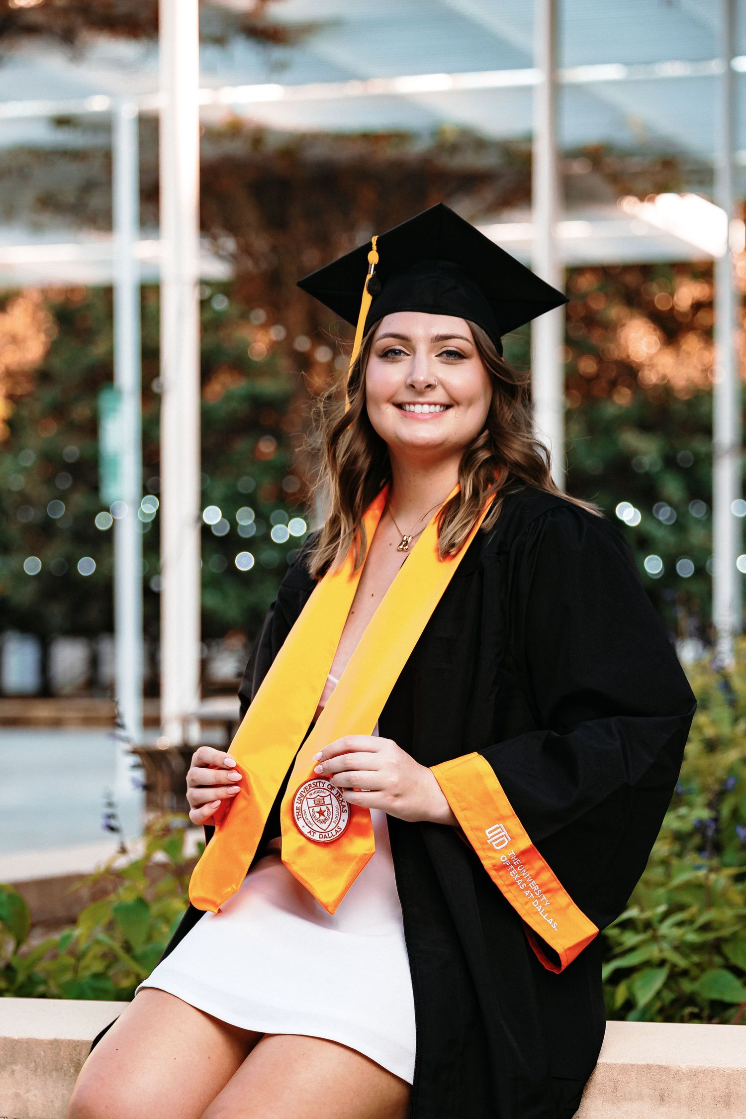 Graduate in black cap and gown with yellow stole sitting outdoors during sunset.