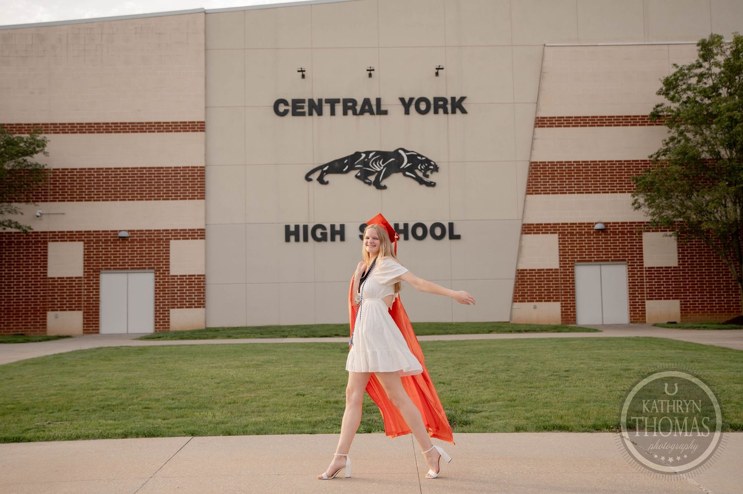 Runner in orange shorts sprints across a grassy field in front of a brick high school building on a sunny day.