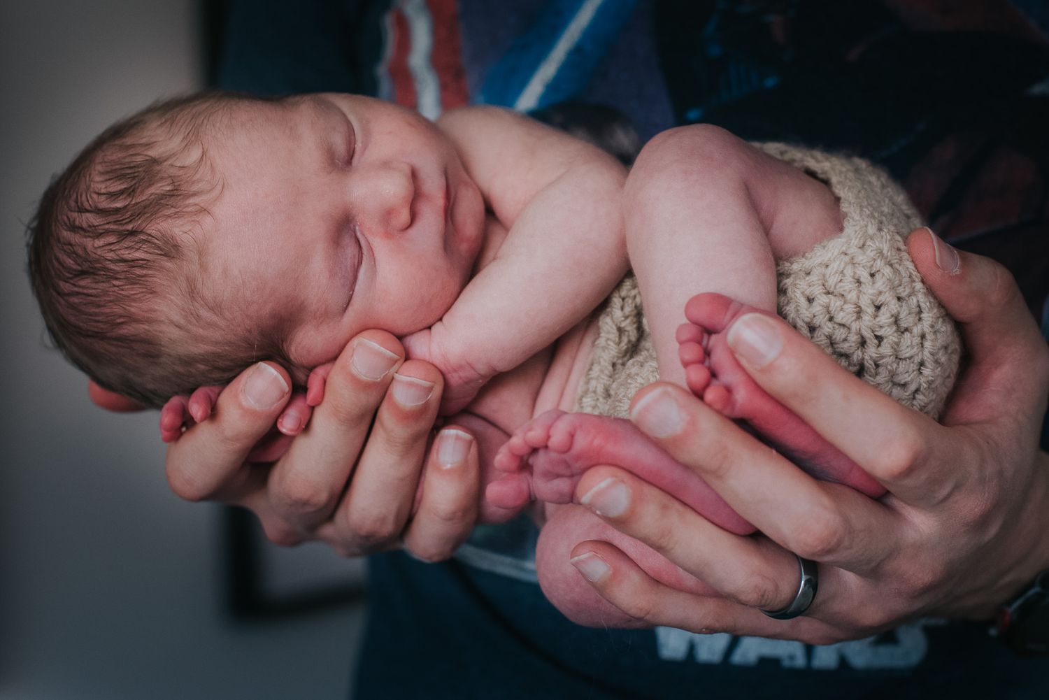A sleeping newborn baby wearing a knit wrap is gently cradled in supportive hands.