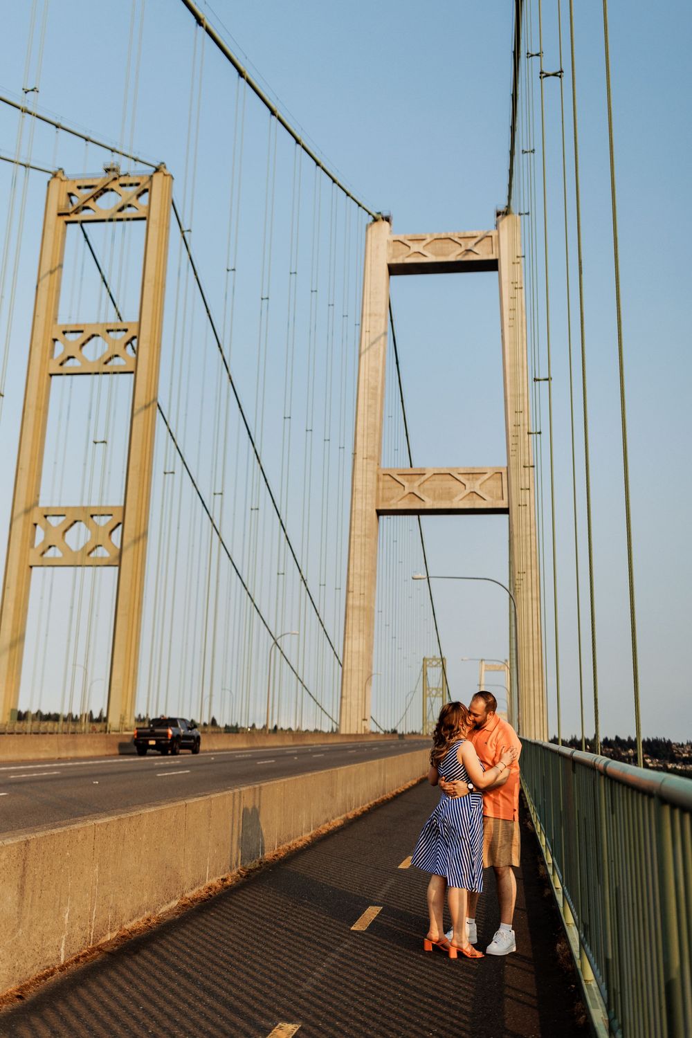 People in golden sunset light stand on the Tacoma Narrows Bridge walkway with suspension cables in the background.