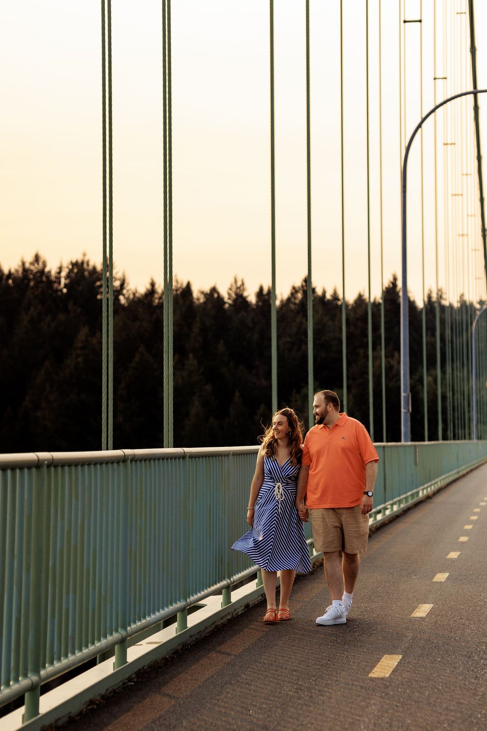 A couple walking together on a bridge at sunset, with the woman wearing a blue dress and man in orange shirt.