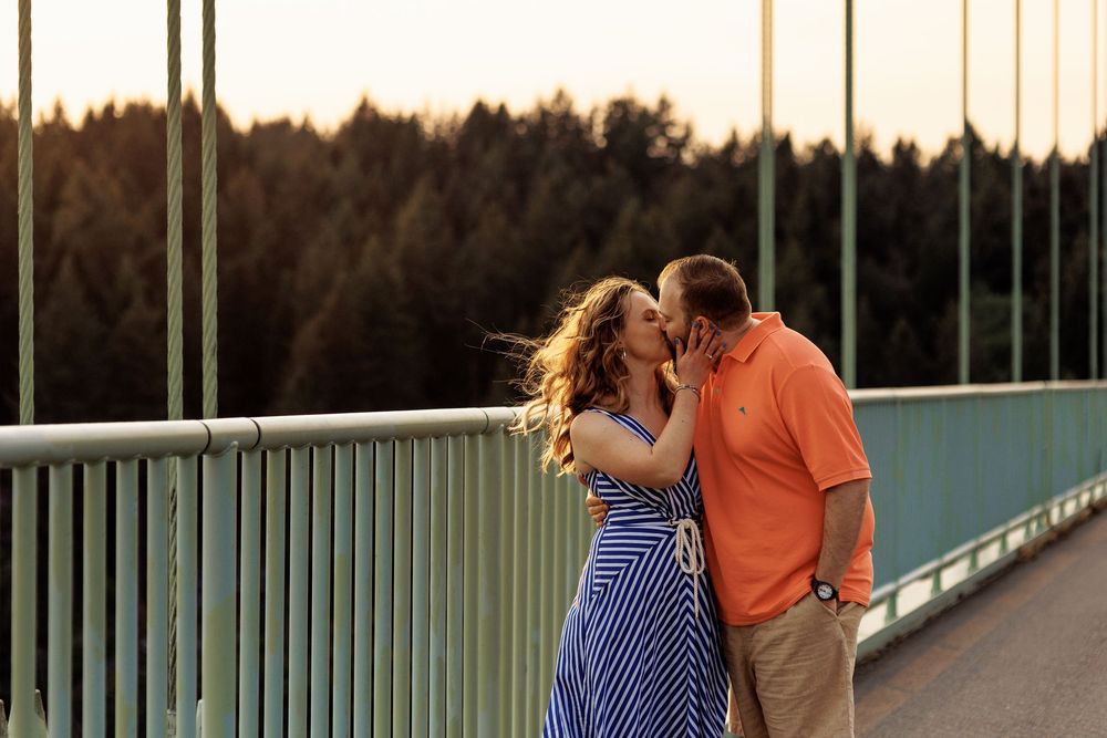 A couple share an intimate moment during golden hour on a bridge with a forested backdrop at sunset.