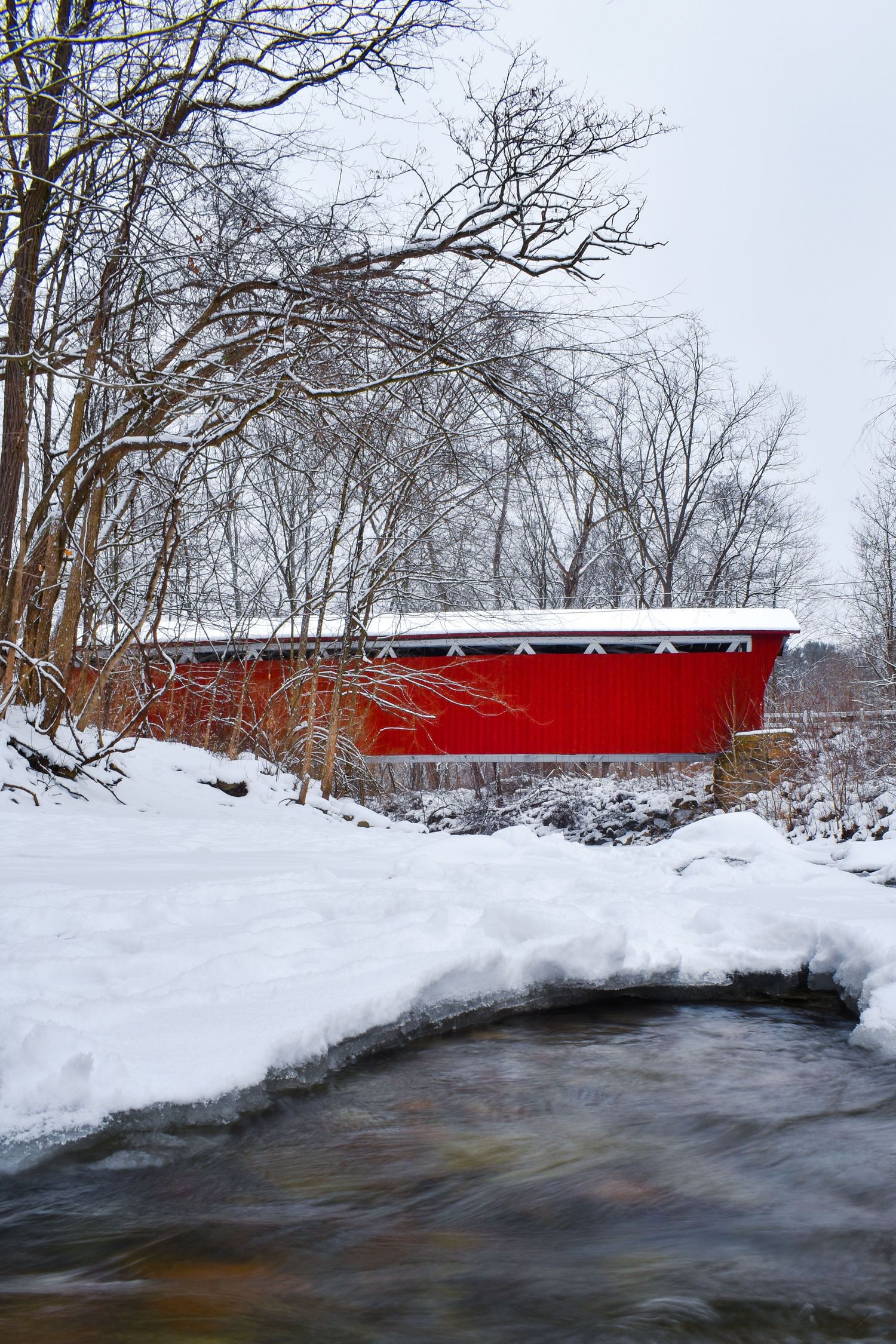 Red covered bridge spans a snow-lined river during winter with bare trees in the background.