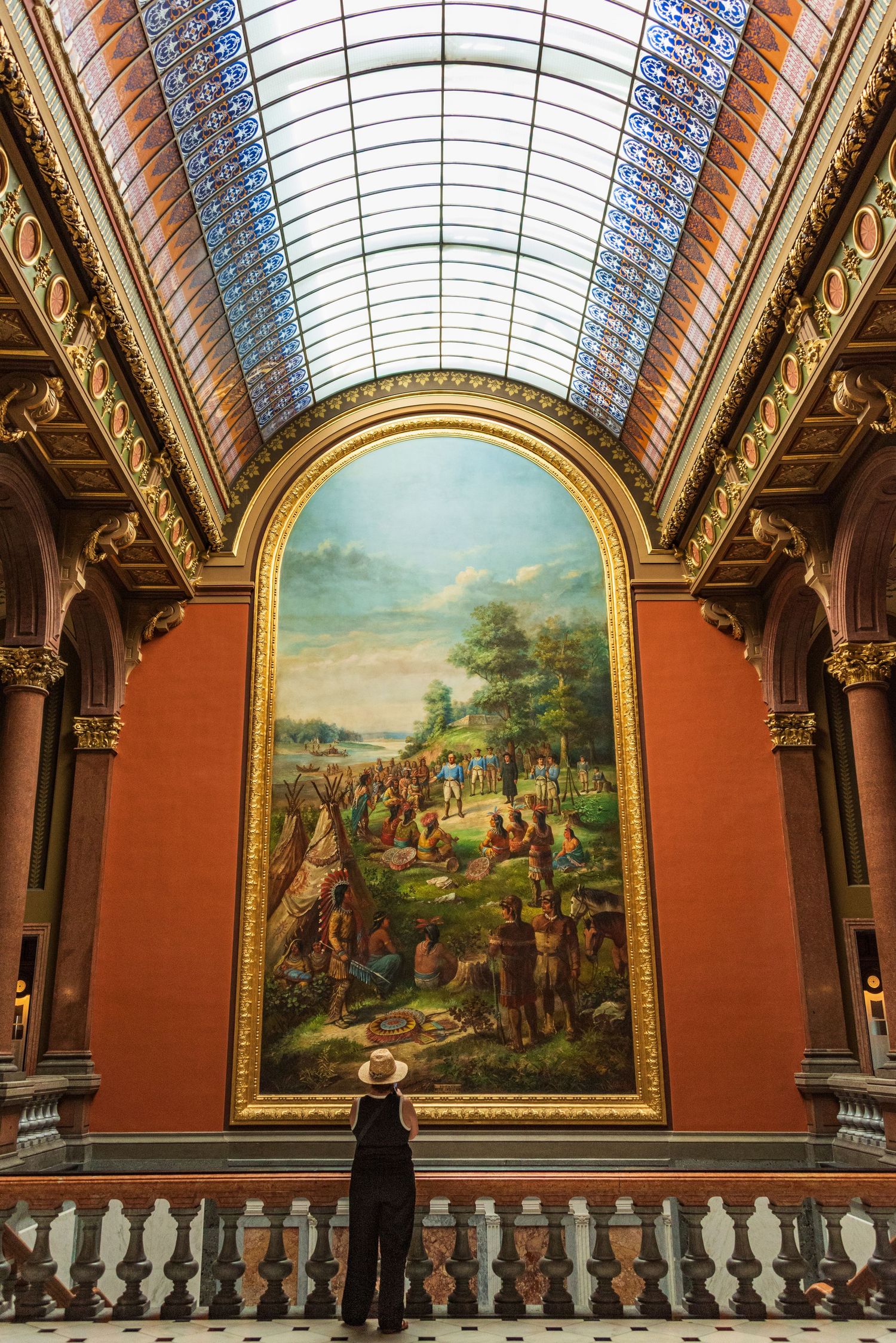 Ornate museum gallery with vaulted glass ceiling and large historical paintings on terracotta walls.