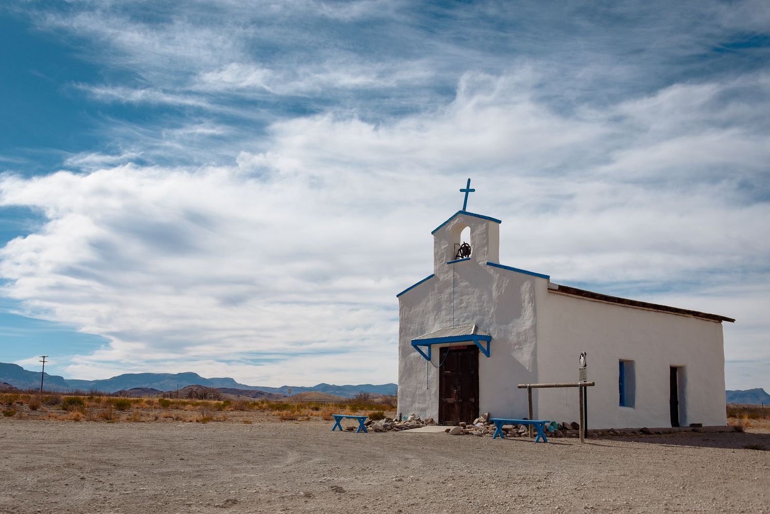 Small white adobe church stands solitary against a dramatic cloudy sky in a desert landscape.