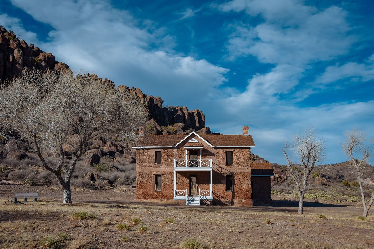 Historic brick ranch house with white porch set against dramatic rocky cliffs in arid landscape.