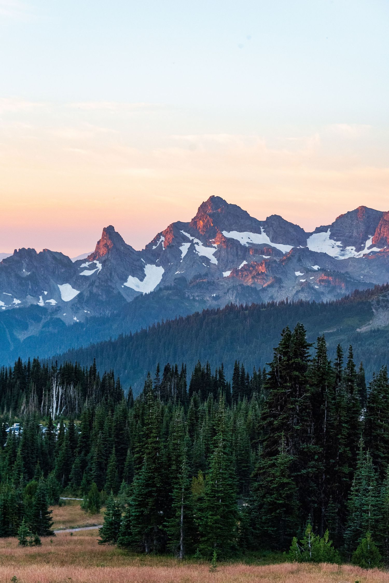 Snow-dusted mountain peaks rise above evergreen forest at sunset in a dramatic alpine landscape.