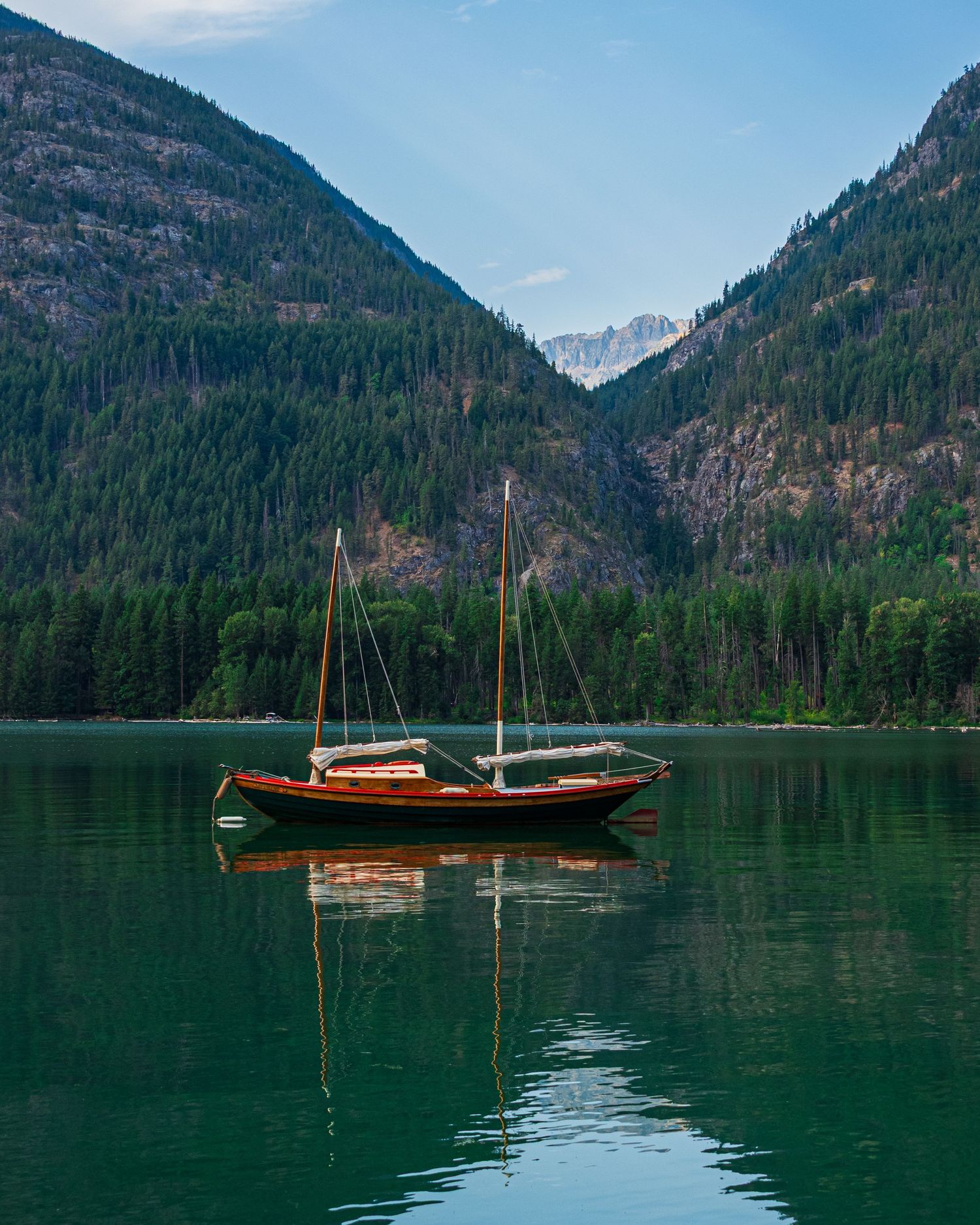 Two wooden sailboats float on calm mountain lake surrounded by forested peaks.
