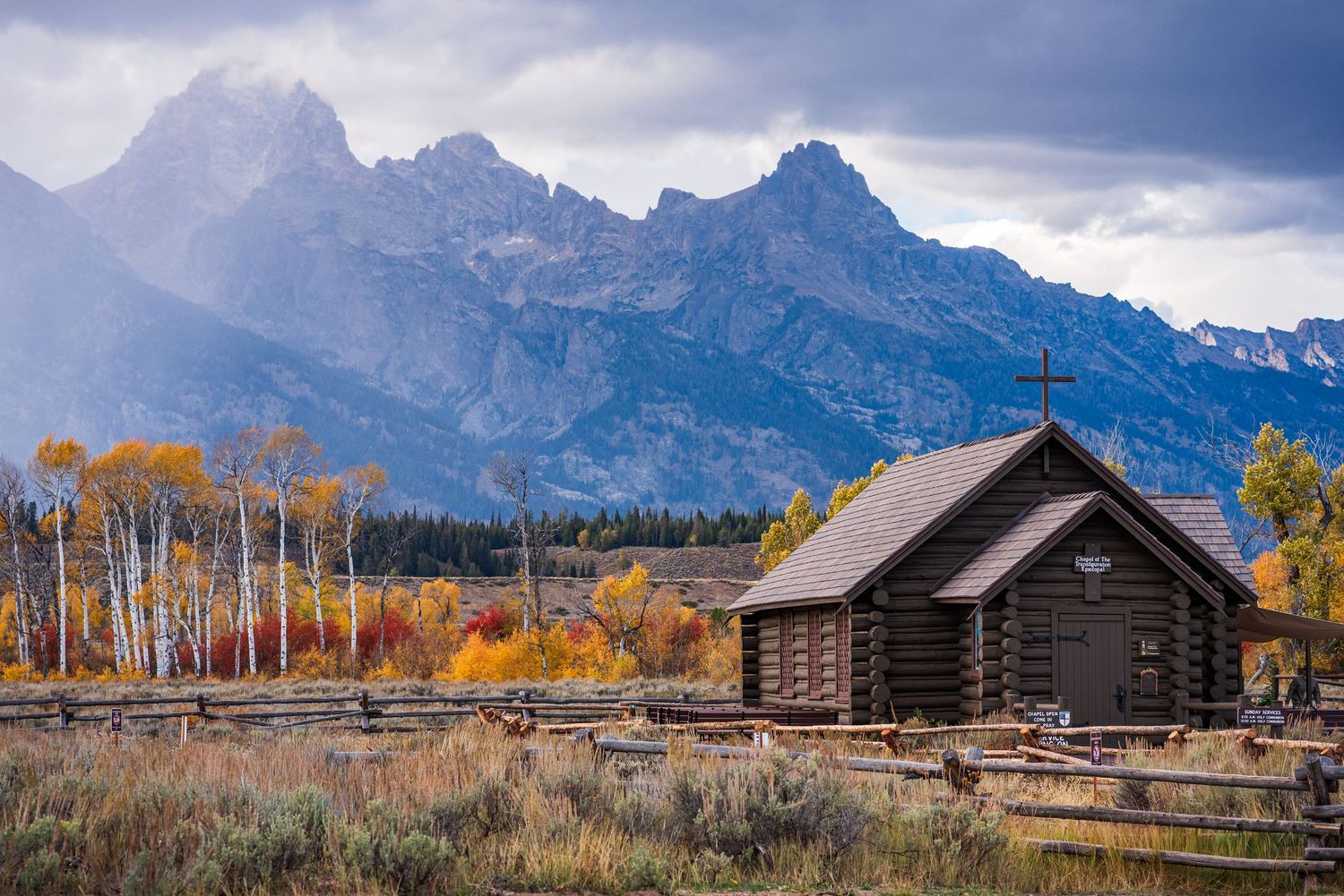 Historic log cabins sit beneath towering mountain peaks with fall colors in Grand Teton National Park.