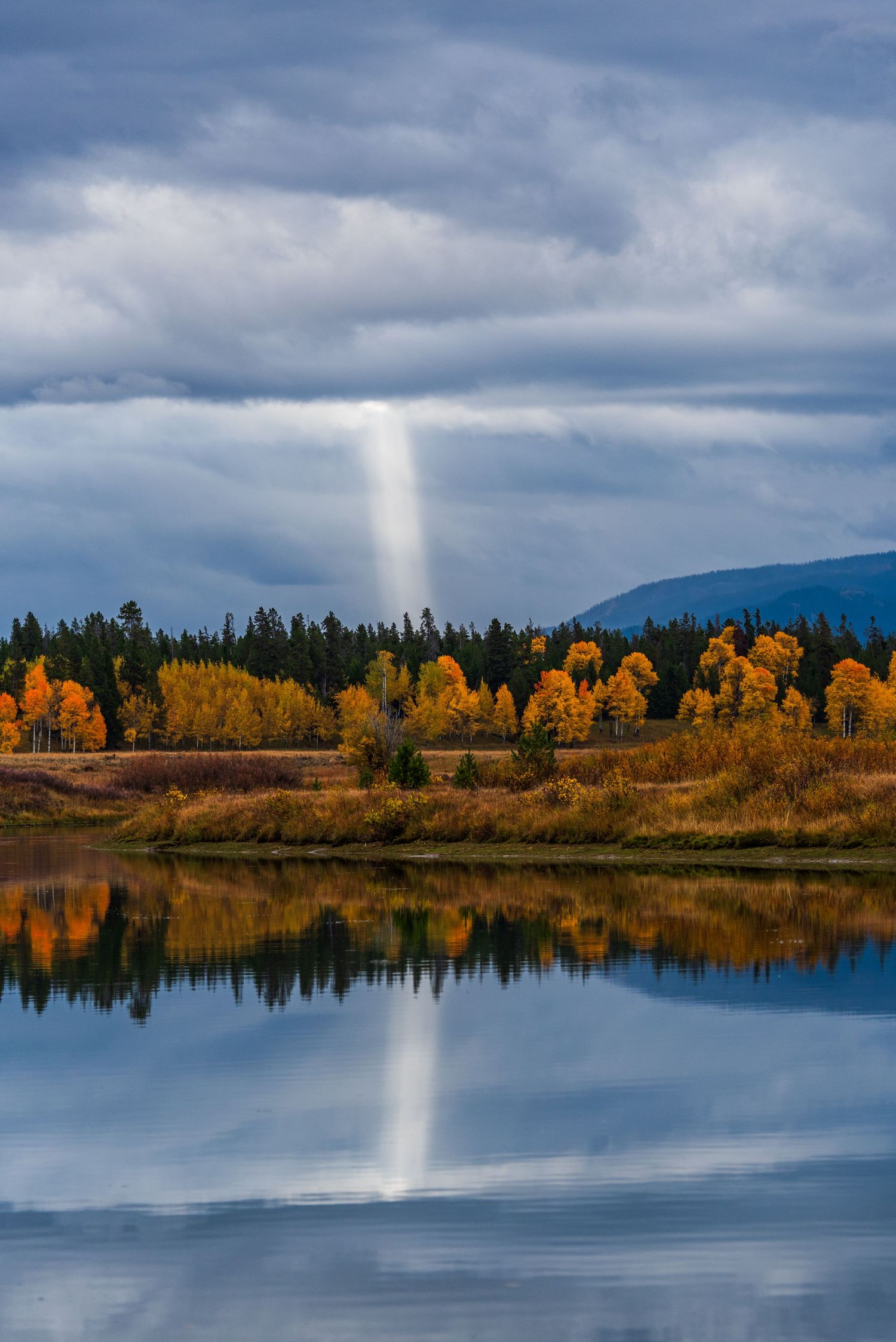 Autumn trees with golden foliage reflect in a calm mountain lake under cloudy skies.