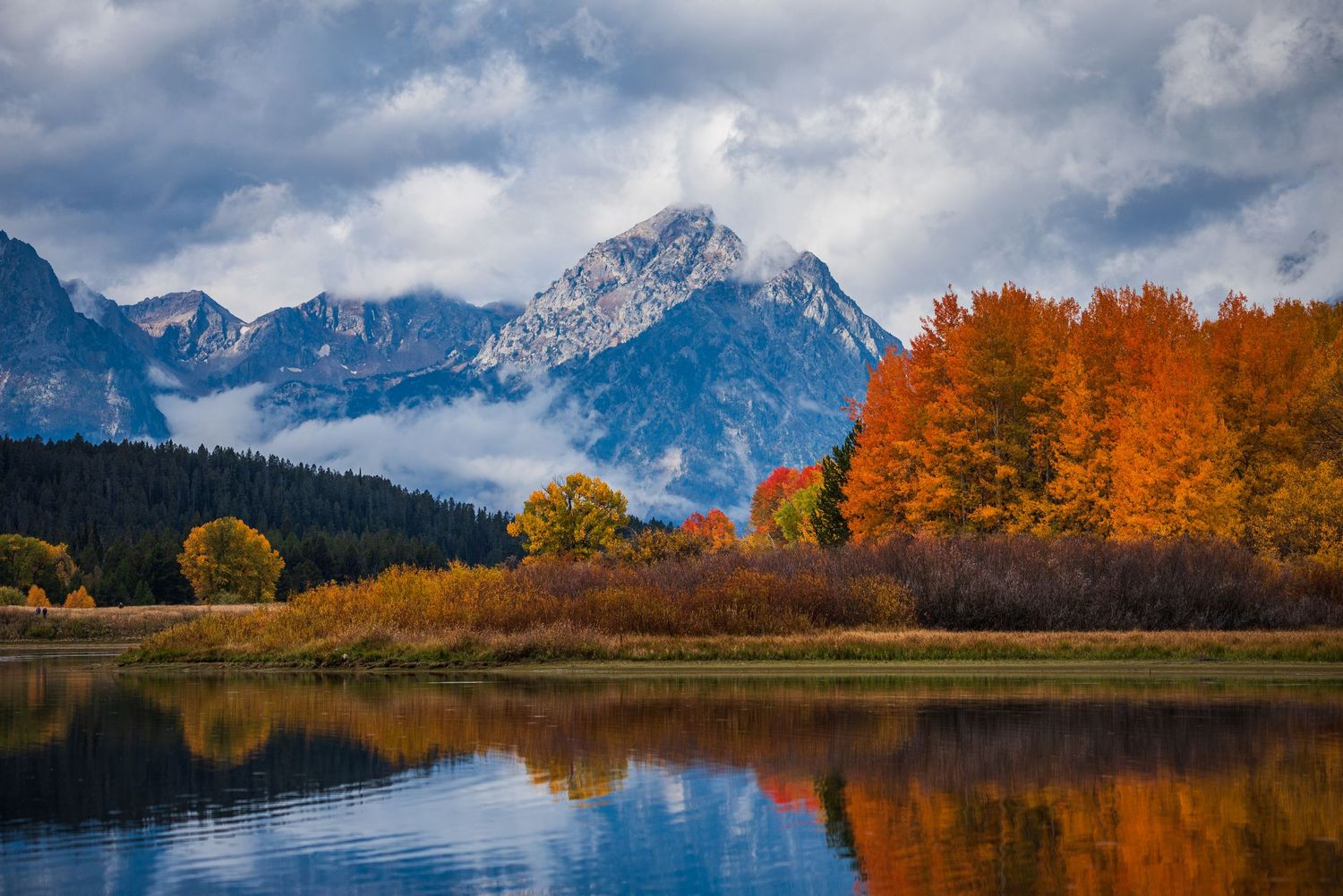 Snow-capped mountain peaks reflect in calm lake waters surrounded by vibrant fall foliage.