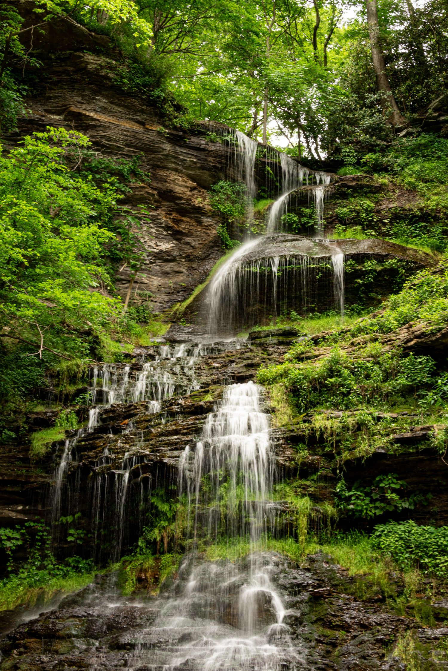 Multi-tiered waterfall cascades down moss-covered rocks in a lush green forest setting.
