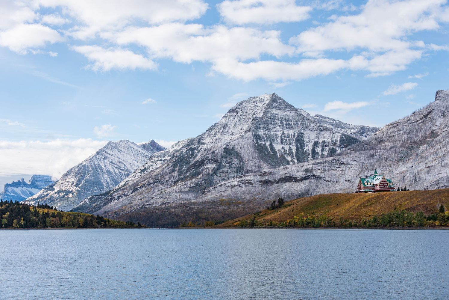 Snow-capped mountain peaks rise above a serene lake under blue skies with white clouds.