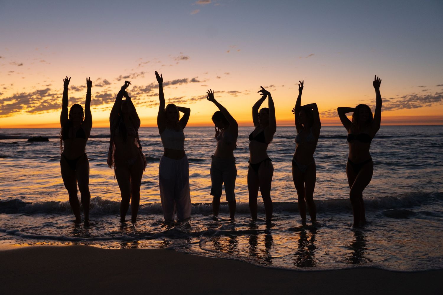 A group of silhouetted people raise their arms during sunset on a beach, creating a joyful and celebratory scene.