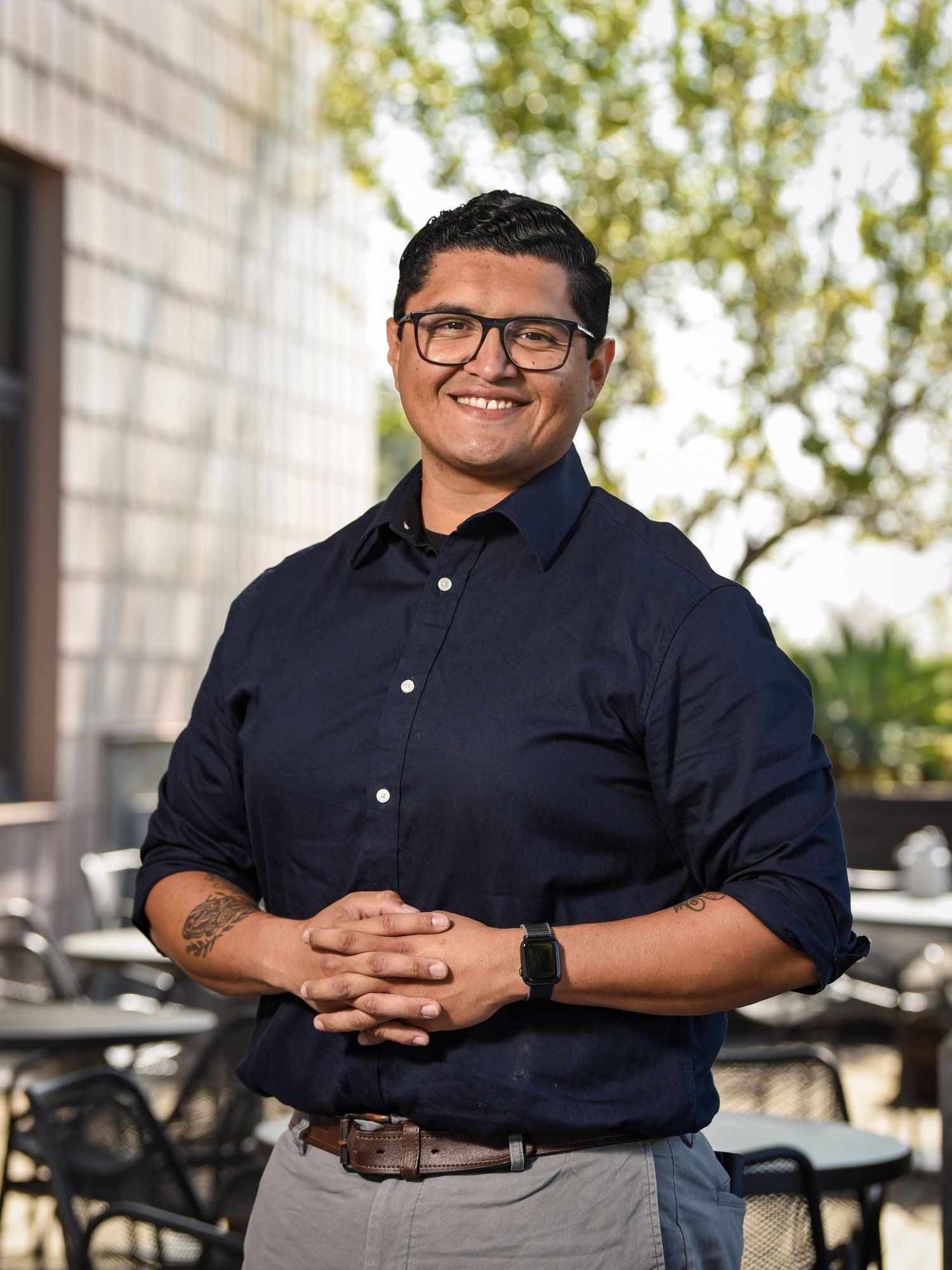 Person in navy button-up shirt and gray pants standing outdoors by patio tables.