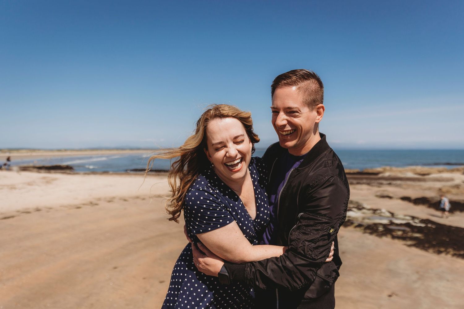 A couple shares a laughing embrace on a coastal cliff overlooking the ocean.