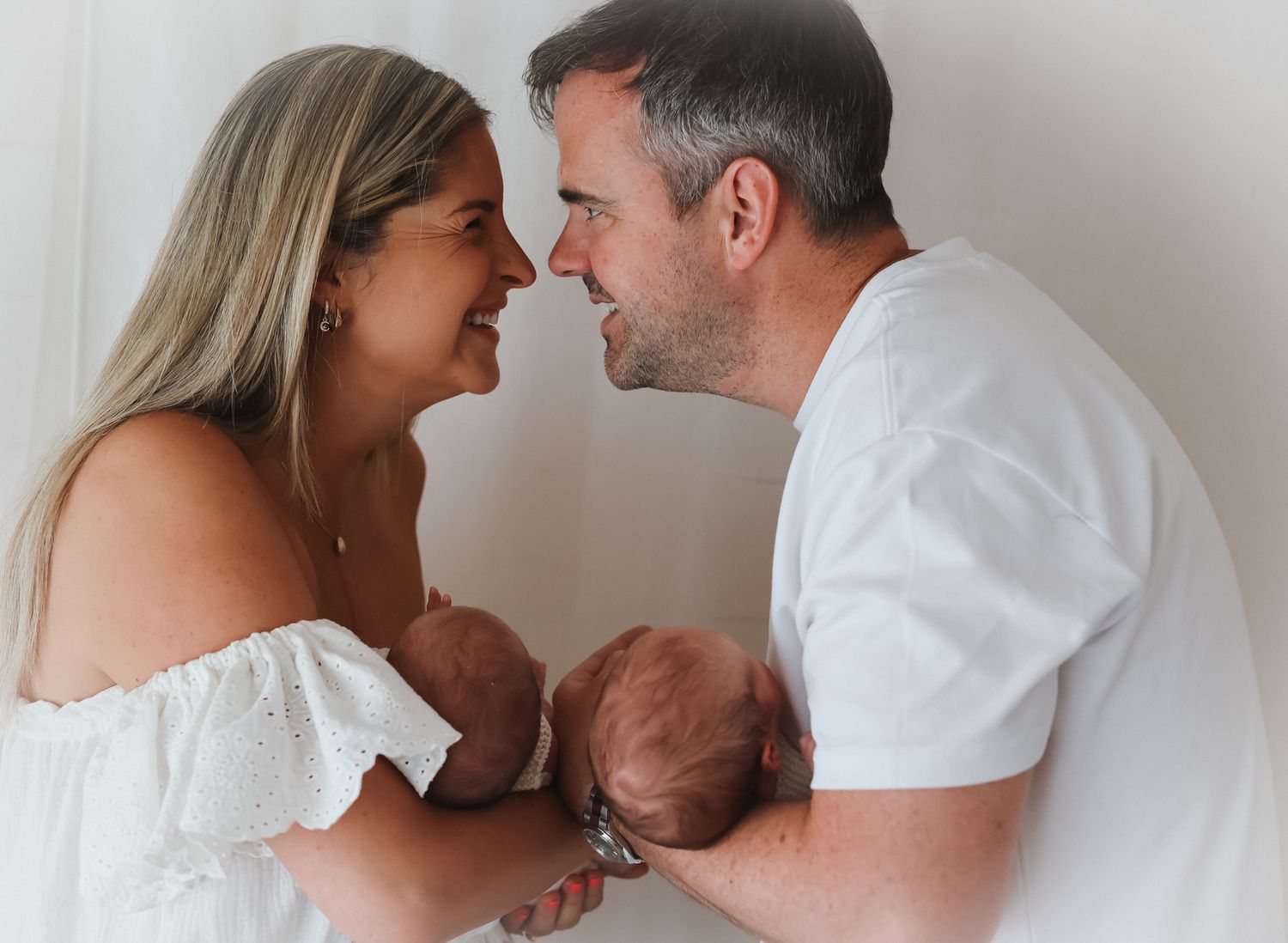 A tender family moment as parents share an intimate close-up while holding their newborn baby against a neutral background.