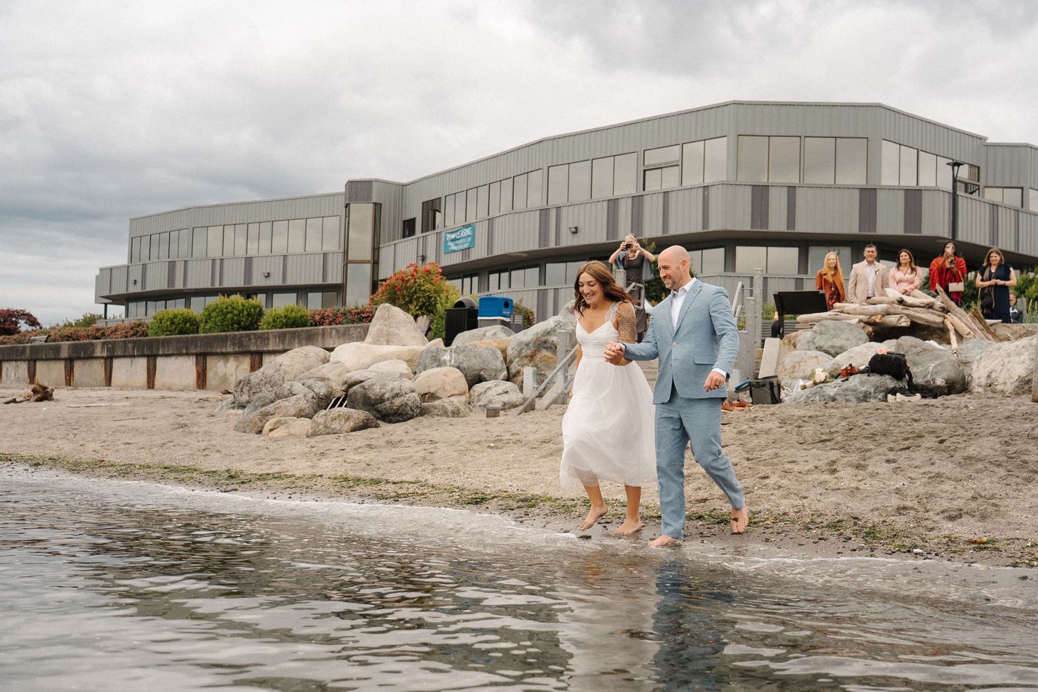 A couple in wedding attire walks along the shoreline near a modern beachfront venue on an overcast day.